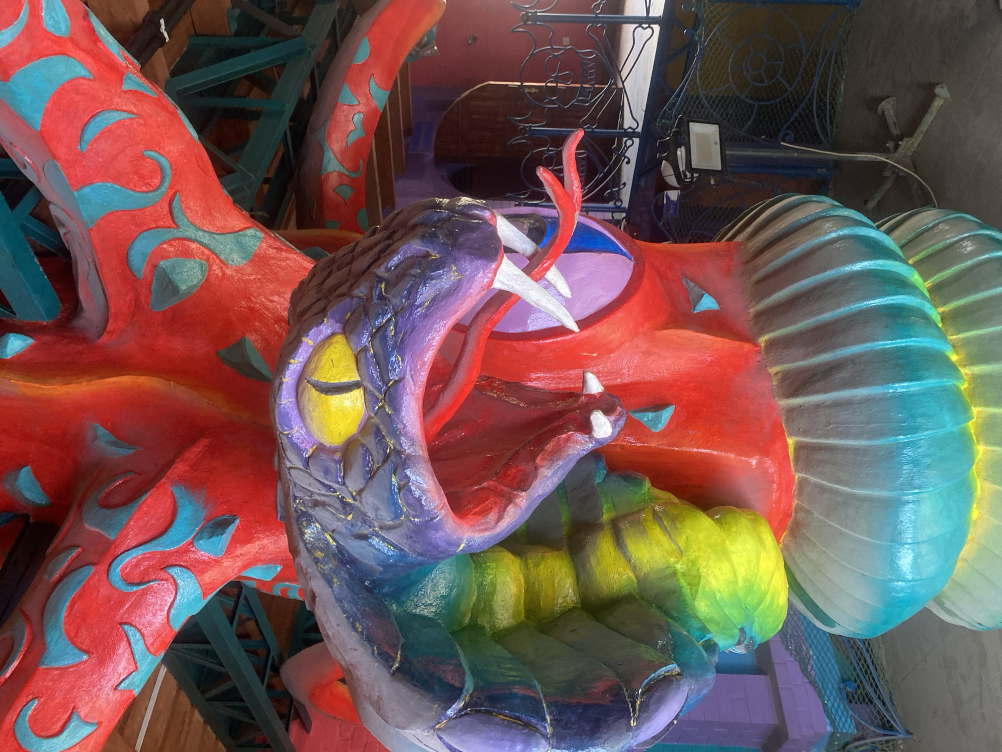 The image displays a vibrant, multi-colored sculpture of a fantastical serpent or dragon. The creature's head is prominently featured, depicted with an aggressively wide-open mouth, revealing a red interior, two large white fangs on the upper jaw, and smaller fangs on the lower jaw. A forked red tongue extends from its mouth. The head is primarily purple, with striking yellow eyes and dark pupils, and its lower jaw and neck feature gradients of green, blue, and purple.
The main body of the sculpture is a brilliant red, adorned with intricate, swirling patterns in teal or light blue. Further along the body, towards what appears to be the tail, the sculpture transitions into segmented, ridged sections that display a gradient of colors from lime green to light blue, suggesting a textured, scaly, or shell-like surface.
The setting appears to be an indoor or covered space, possibly an exhibit, theme park attraction, or cultural display. In the background, dark green or teal metal structural beams are visible. An ornate, dark metal grate or railing with decorative swirls is also present, to which a black floodlight is attached. Other background elements include reddish-brown walls and darker, possibly purple or grey, floor or wall areas. The lighting suggests artificial illumination. There are no people present, and no visible text in the image. The overall impression is one of energetic, fantastical artistry.
