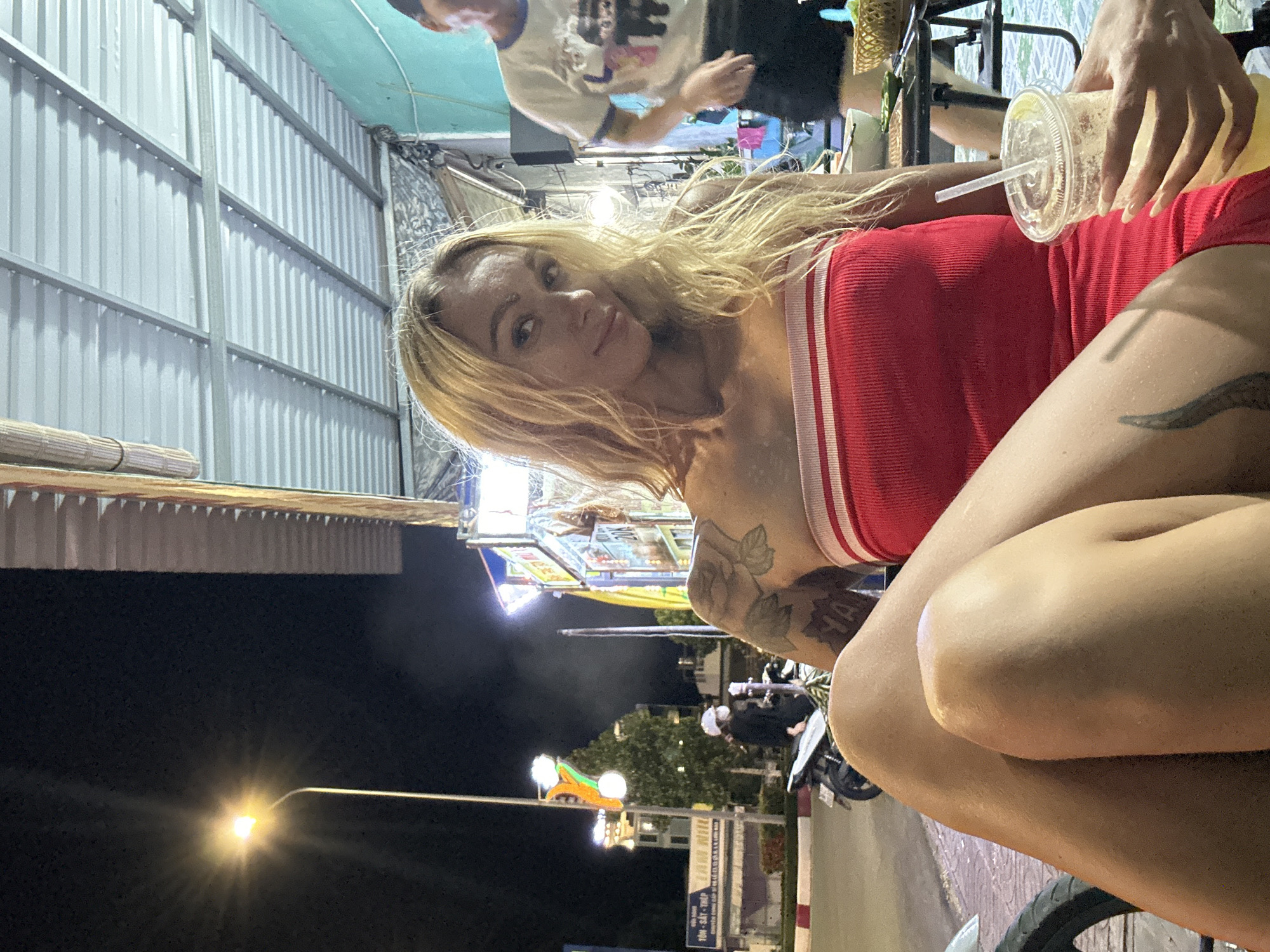 An exterior shot shows a woman with blonde hair and tattoos on her arm, wearing a red strapless dress and holding a drink. She is seated outdoors at night, with a streetlight and a building facade visible in the background. The scene appears to be a street-side establishment in Phu Thuy Ward, Vietnam, as indicated by signage. A person in a white t-shirt and black shorts is also visible in the background.