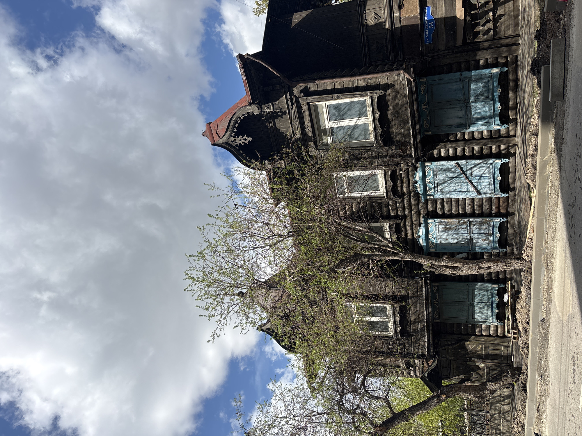 The image depicts a two-story wooden house in Krasnoyarsk, Russia. The house is dark brown, with intricately carved details on the eaves and window frames. The windows on the upper floor are white, while the lower-floor windows have blue shutters. A tree with budding green leaves is in front of the house, partially obscuring the view. A blue street sign labeled "ул. Аллы Лебедевой, 34" (Alla Lebedeva Street, 34) is attached to the house. The sky is visible in the background, with a mix of blue and white, cloudy skies. A portion of the sidewalk and street can be seen on the right side of the frame.