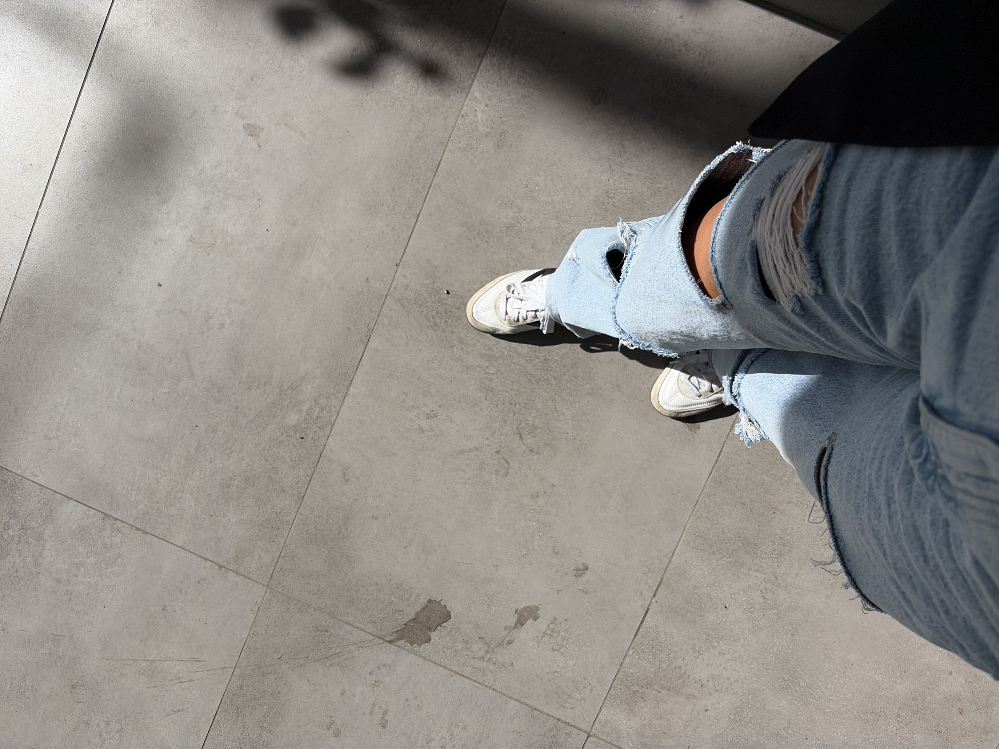 The image shows a person's legs from the knees down, wearing ripped light-wash denim jeans and white sneakers. The person is standing on a light gray tiled outdoor surface, possibly a sidewalk or patio. Shadows from overhead foliage are cast across the tiles, indicating it is daytime. There is no indication of the location being Moscow, Russia. The scene is neutral, with no specific action or interaction depicted.