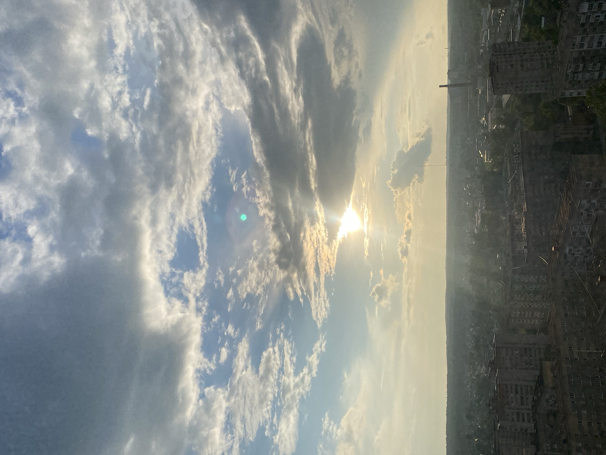 The image depicts a panoramic view of the city of Izhevsk, Russia, dominated by a dramatic sky filled with a mixture of dark and light clouds. The sun is partially obscured by these clouds, casting a soft, diffused light over the cityscape below. The city itself appears to be composed of a dense collection of apartment buildings and other structures, with a noticeable haze or mist hanging over the horizon, obscuring distant details. The overall impression is one of a slightly overcast day with the sun attempting to break through.