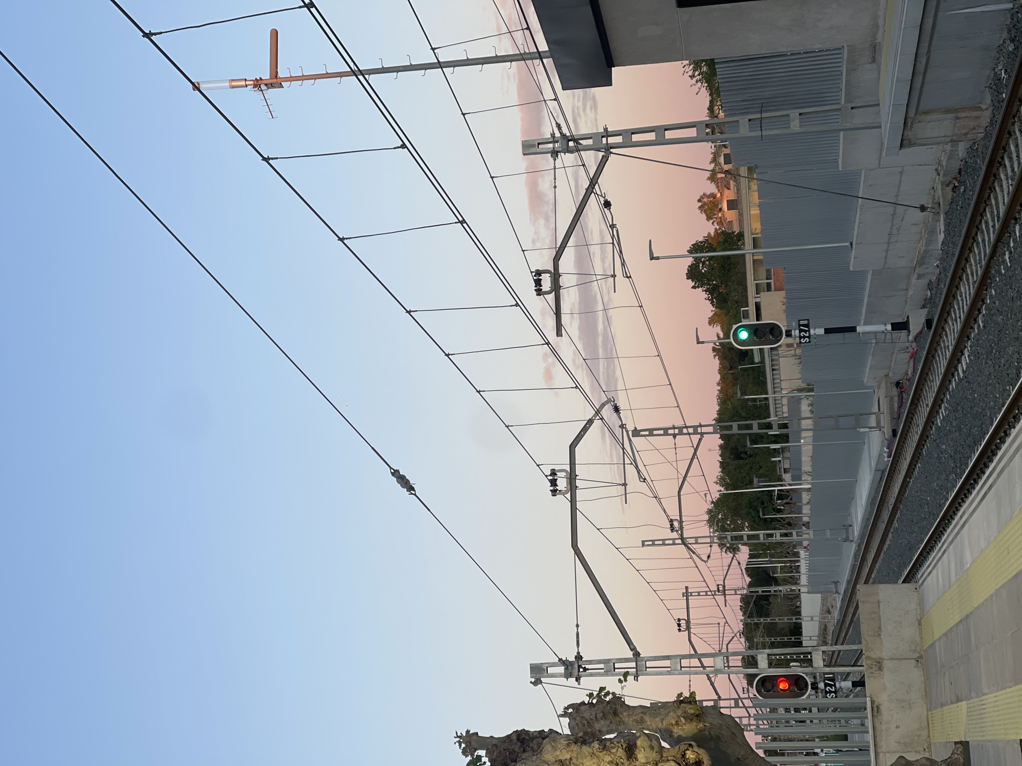 The image depicts a railway scene in Paiporta, Spain. Overhead power lines and supporting structures are prominently visible against a backdrop of a twilight sky with hues of pink and blue. A train track runs along the right side of the image. Train signals with red and green lights are observed along the tracks. A platform edge with yellow tactile paving is present. A grey, slatted wall is seen behind the train tracks. Tree foliage is present near the bottom of the image.