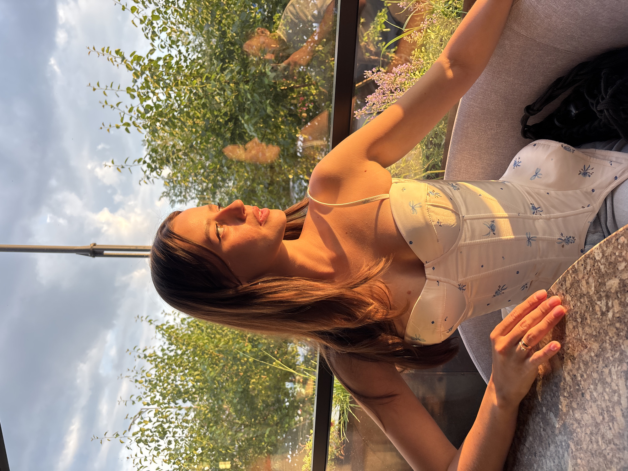 In Krasnoyarsk, Russia, a young woman with long brown hair sits at a table by a window, illuminated by the warm glow of the setting sun. She is wearing a white corset top adorned with a delicate blue floral pattern. Her left hand rests on the table, displaying a ring on her finger. Through the glass window, a reflection of the woman can be seen, along with another person, possibly a man, holding a camera. Green foliage and purple wildflowers are visible outside, suggesting an outdoor seating area. The sky is partly cloudy, with the soft light of the golden hour casting long shadows.