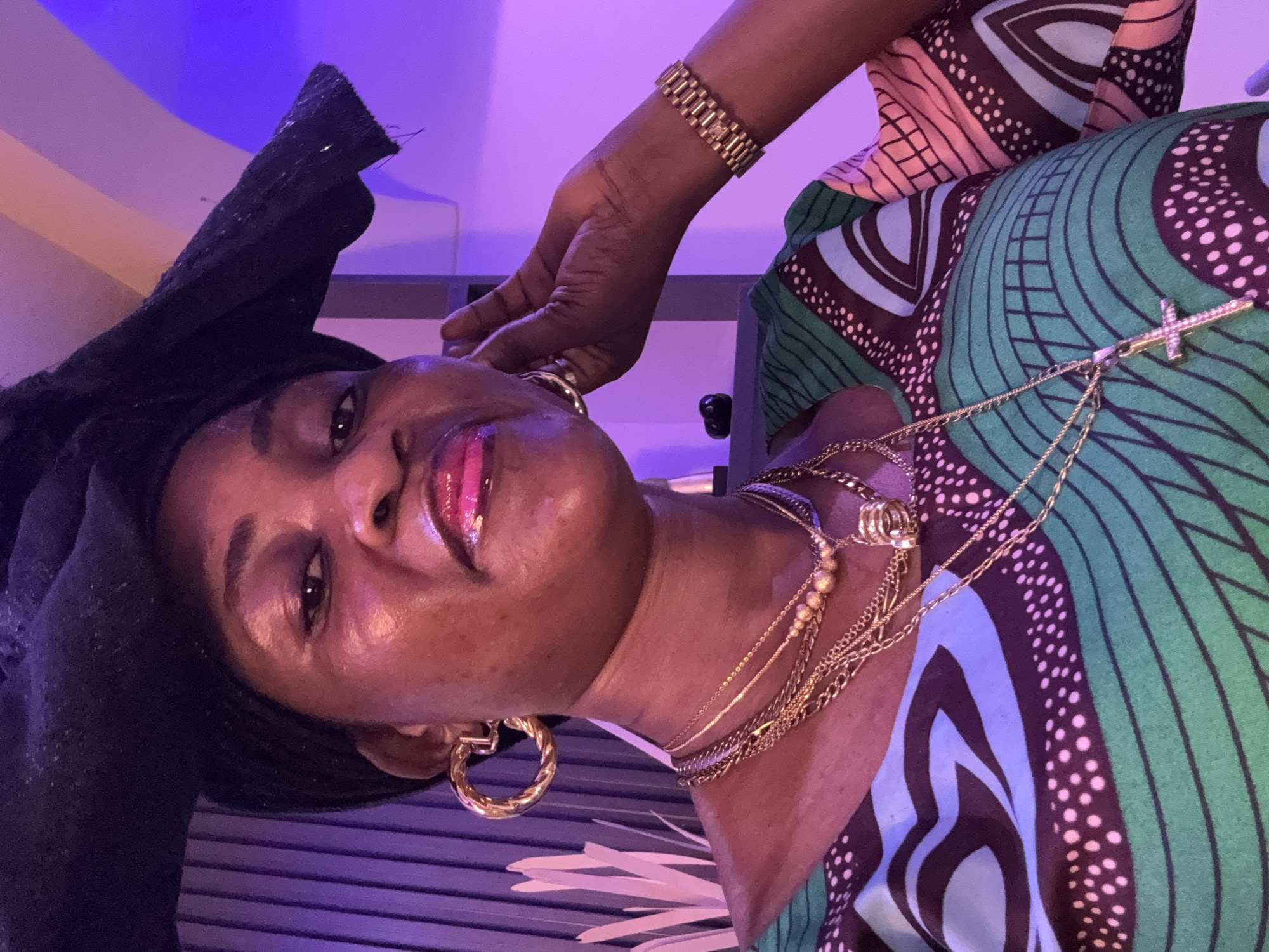A middle-aged Black woman with dark skin and wearing a black headwrap is positioned in the center of the frame, looking directly at the camera. She has bright pink lipstick on and is wearing multiple gold necklaces, large gold hoop earrings, and a gold watch on her left wrist. She is also wearing a colorful green, blue, and pink patterned top. The background is blurred, with vertical blinds on the left and a purple wall on the right. The lighting appears to be artificial, casting a pinkish and purplish hue over the scene. The woman's right hand is raised to her ear. The scene appears to be indoors, possibly in a residence or a social gathering.