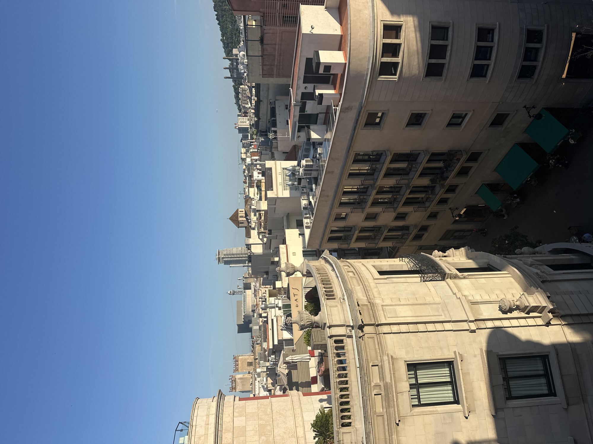 This is an aerial, eye-level, outdoor, daytime, vertical shot of the Barcelona cityscape. The sky is clear and blue, with a few small, distant specks, possibly birds or planes. The foreground features the upper floors and rooftops of several buildings.
On the left, a rounded, light-colored stone building with classical architectural details is prominent. It has large windows with sheer curtains and ornate balconies with metal railings. A rooftop terrace with lounge furniture and umbrellas is visible on this building.
To the right of the stone building is another building with a curved facade, painted in a light beige color. This building has multiple floors with rows of windows, some with balconies and metal railings. Green awnings are extended over storefronts on the ground floor of this building, and patrons are visible seated at tables beneath them.
Further into the background, a dense collection of buildings of varying heights and architectural styles stretches across the horizon. Some buildings are modern, with flat roofs and air conditioning units, while others have pitched roofs and older designs. Distant structures include towers, a dome, and what appear to be industrial chimneys on a hill in the far background. The overall impression is of a dense, urban environment bathed in warm sunlight.