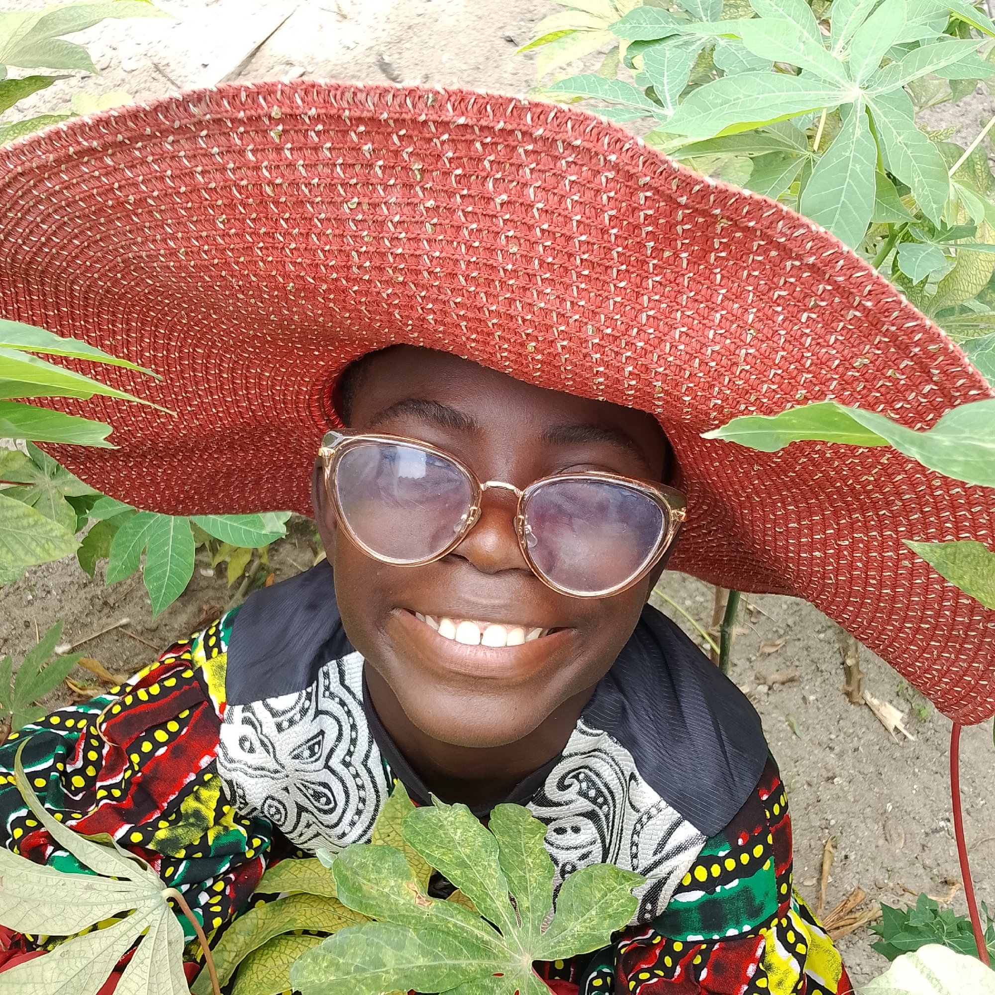 A young person wearing a wide-brimmed red hat and glasses is smiling at the camera. They are dressed in a colorful, patterned shirt with black accents. The person is surrounded by green foliage and appears to be outdoors in a rural or garden setting. The ground is sandy or earthy. This scene is likely in Ado, Nigeria, based on the implied context.