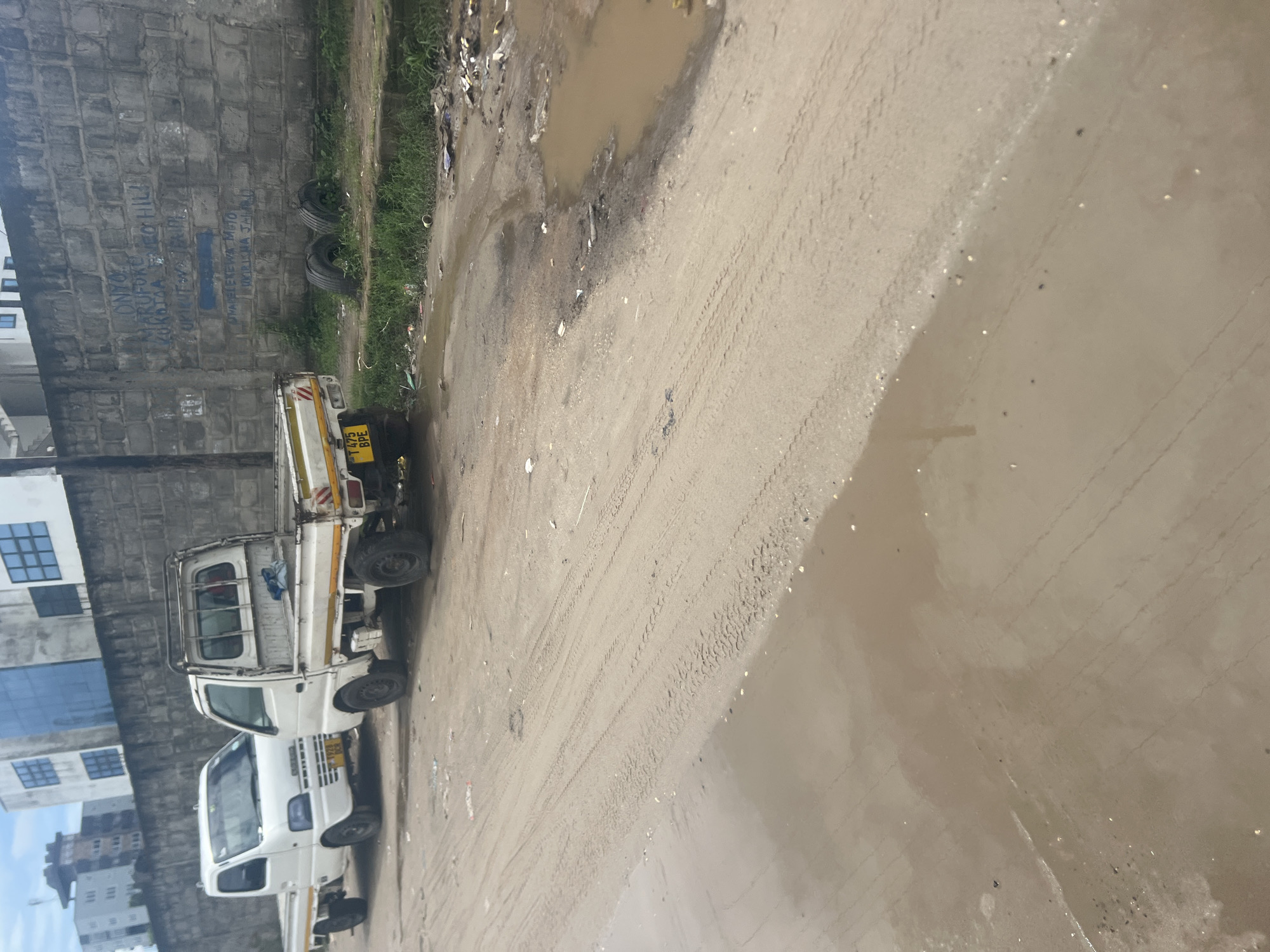The image depicts a street scene in Dar es-Salaam, Tanzania. A white truck is parked on the side of a dirt road. There is standing water in the road and trash along the roadside. A concrete block wall lines the left side of the frame. Graffiti is visible on the wall. Two tires are leaning against the wall. A building with windows is visible behind the wall.