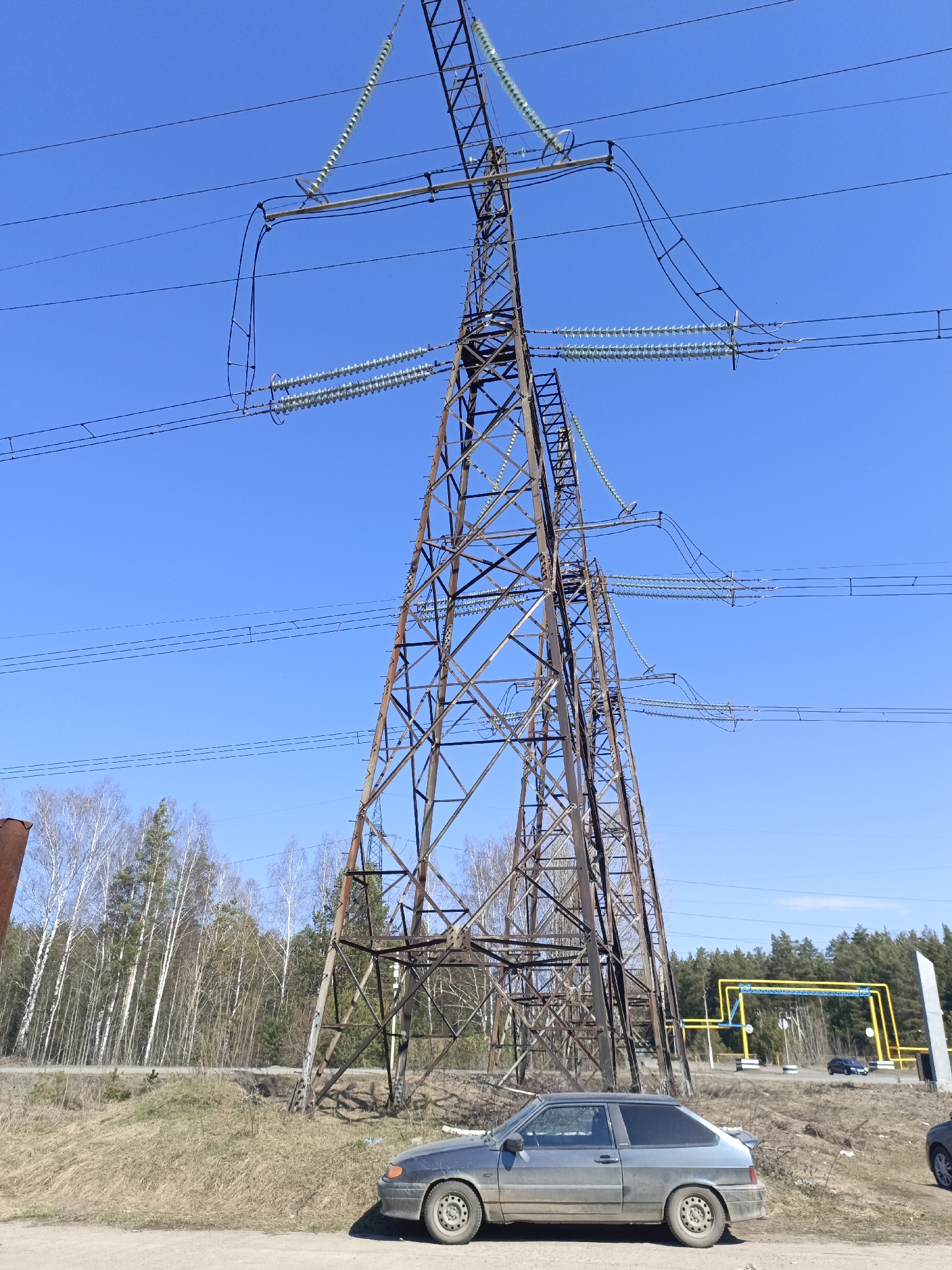 The image captures a rural or semi-urban landscape under a clear, bright blue sky, likely in Noviy, Russia. The dominant features are a large, weathered lattice-steel electricity transmission pylon and its associated high-voltage power lines. The pylon, showing signs of rust, supports multiple conductors on its crossarms, which are insulated by long strings of disc insulators. Another similar pylon is partially visible in the mid-ground.
In the foreground, a dusty, grey two-door hatchback car, resembling a Lada Samara, is parked on a dirt and dry grass shoulder. Behind the car and to the left, a forest composed mainly of bare deciduous trees (birches) and some evergreens stretches across the horizon. To the right, a denser coniferous forest is visible.
A notable man-made feature in the mid-ground is a yellow gas pipeline structure with blue support beams, crossing over a paved road. A distant car can be seen on this road. The general impression is one of utility infrastructure intersecting with nature. The time of day is daytime, indicated by the bright sunlight and lack of long shadows, and the bare trees suggest an early spring season. No people or visible text are present.