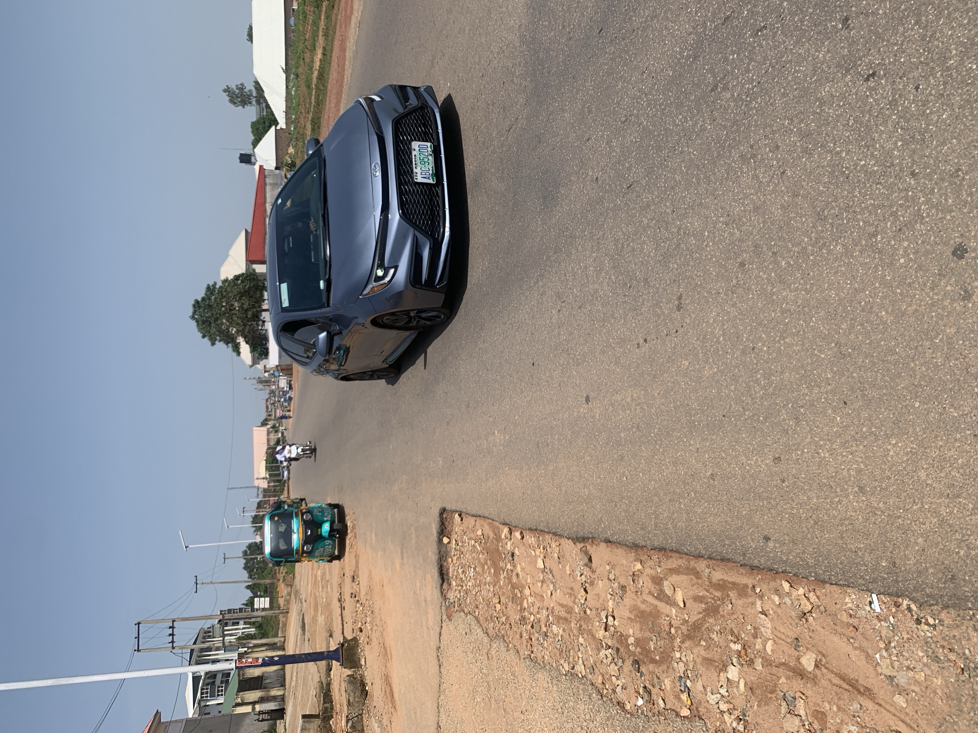 The image depicts a street scene in Jos, Nigeria, under a clear sky. A gray Toyota sedan with a Nigerian license plate ABC-95200 is driving on the right side of the asphalt road. To the left of the road, a blue and green auto-rickshaw is parked. Further in the background, a person is riding a motorcycle. The road shoulders are unpaved and eroded in places. Buildings line the street on the left, with utility poles and power lines visible. The overall environment suggests an urban or semi-urban setting with ongoing infrastructure development or maintenance issues.