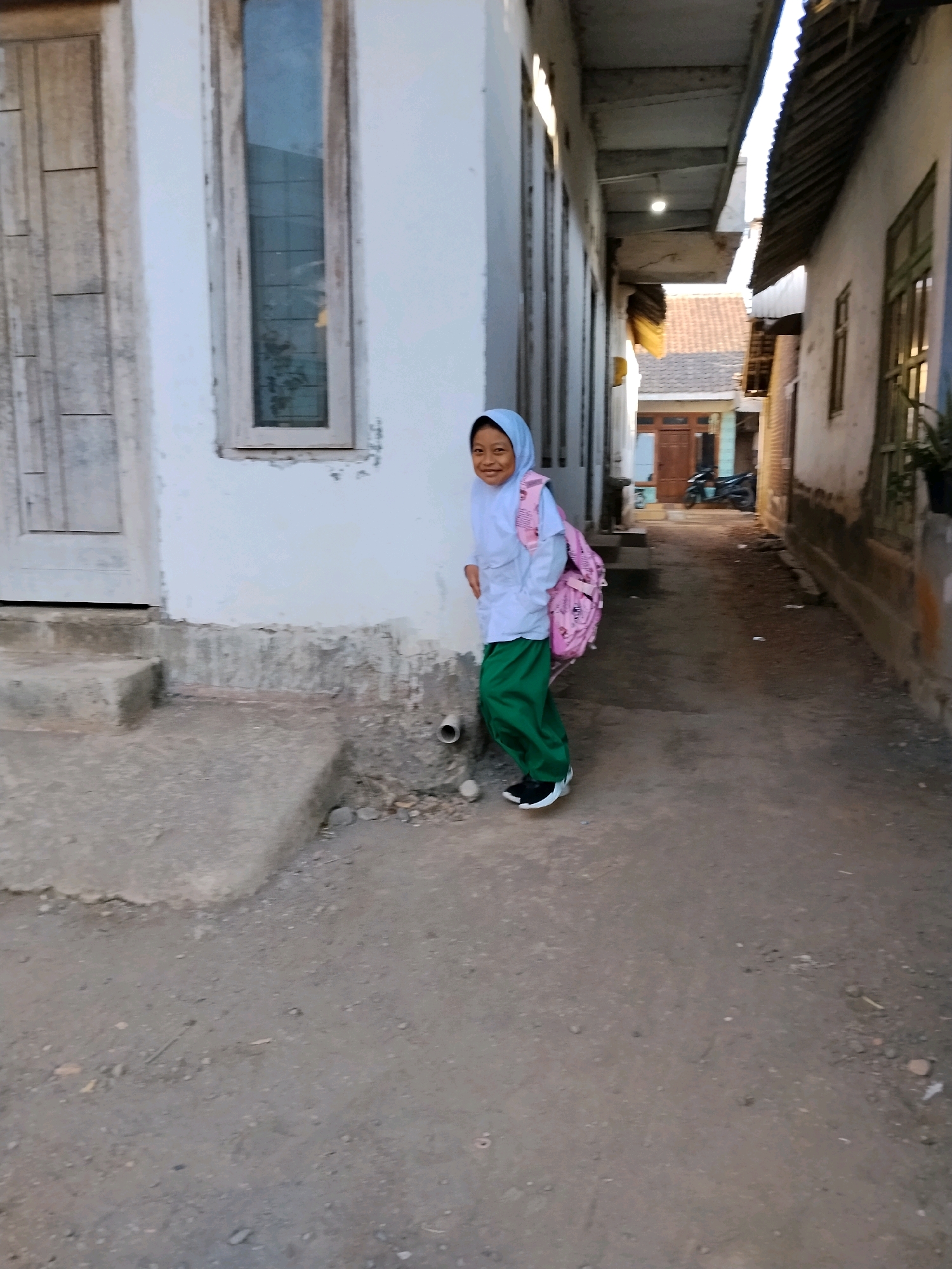 A young girl wearing a light blue hijab, white shirt, and green pants stands on a dirt path next to a white building. She has a pink backpack on her shoulders. The building has a wooden door and a tall, narrow window. To the right of the girl, the path leads between two buildings with corrugated metal roofs, suggesting an alleyway. In the background, there's a motorcycle parked in front of a wooden door. The overall scene depicts a residential area in Panggungan, Indonesia.