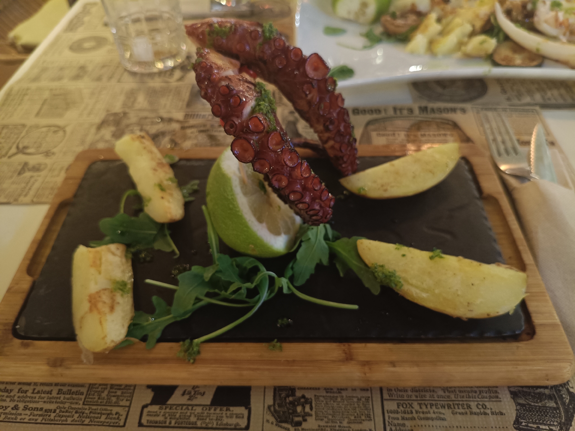 A close-up, eye-level shot captures a platter of food on a dark slate board resting on a wooden tray. The food consists of several pieces of cooked octopus, a wedge of lime, and four wedges of potato. The octopus tentacles are dark red, glistening, and appear to have a green sauce drizzled on them. The potato wedges are golden brown and sprinkled with herbs. Arugula greens are scattered around the food. The background shows a table covered with newspaper-like placemats, a glass of water, and other out-of-focus food dishes. The setting appears to be a restaurant in San Gabriel, Spain.