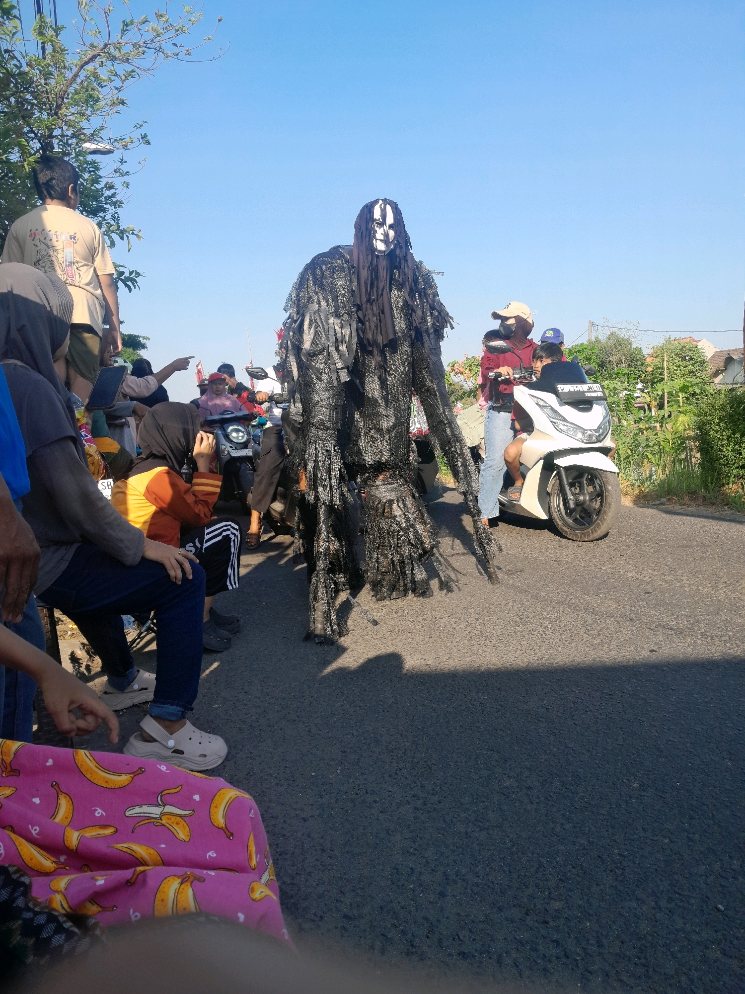 The image shows a street scene in Terung Kulon, Indonesia, under a clear blue sky. In the foreground, a person in a black, ragged costume with a white, painted mask, resembling a figure from folklore or a parade, stands center frame. To the right, two people are on a white scooter. The person in front is wearing a maroon top, blue jeans, and a beige cap, while the person behind is wearing a dark shirt and a blue cap. The scooter has a license plate that appears to read "A 7753 AED".
Several other people are visible in the background. To the left, a person wearing a light brown t-shirt stands near a tree with green leaves. Next to them, a person in a grey hijab and dark clothing is seated. Further back, a group of people are gathered, some appearing to be watching the event. The street is paved, and there are houses and vegetation visible in the distance, indicating a semi-urban or rural setting. A pink cloth with a banana pattern is visible in the bottom left corner of the image. The overall atmosphere suggests a local event or parade.