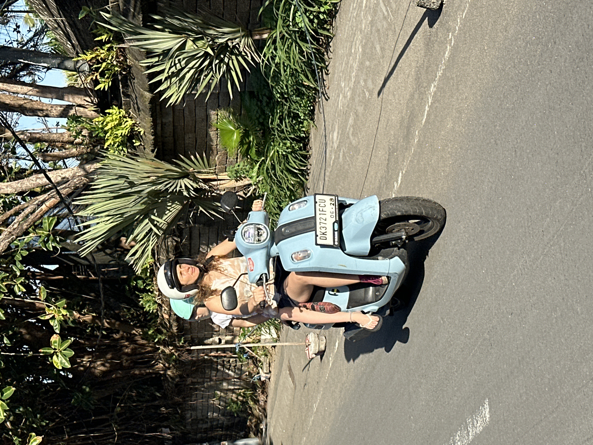 In North Kuta, Indonesia, a light blue Yamaha scooter with two people on board travels on an asphalt road. The driver, a woman wearing a white helmet and a sleeveless top, operates the scooter. Behind her, a passenger with a teal helmet is seated. The scooter displays a license plate "DK 3721 FCU". The scene is bathed in sunlight, casting shadows on the road. Lush green foliage and a stone wall line the right side of the road, suggesting a tropical environment. The scooter is in motion, indicated by the blurred background and the woman's grip on the handlebars.