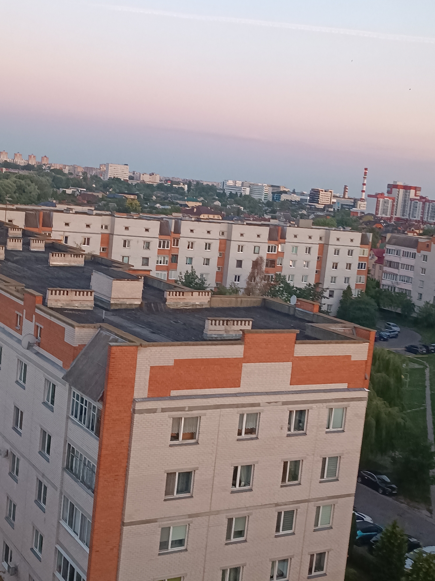 The image depicts an aerial view of a residential area in the city of Brest, Belarus, at dusk. The foreground features the rooftops of multi-story apartment buildings with dark grey roofing material. Some buildings have prominent orange brick accents on their facades and around the chimneys. The buildings are characterized by numerous windows, many with white frames. In the middle ground, more apartment buildings are visible, some with lighter colored facades and orange decorative elements. A tall industrial chimney with red and white stripes stands in the distance, suggesting an industrial zone or power plant on the outskirts of the city. The background shows a sprawling urban landscape with more buildings, trees, and a hazy sky with soft pink and blue hues. Several cars are parked along a road in the lower right portion of the image, and a few birds are visible in the sky. The overall impression is one of a typical urban environment in the evening.