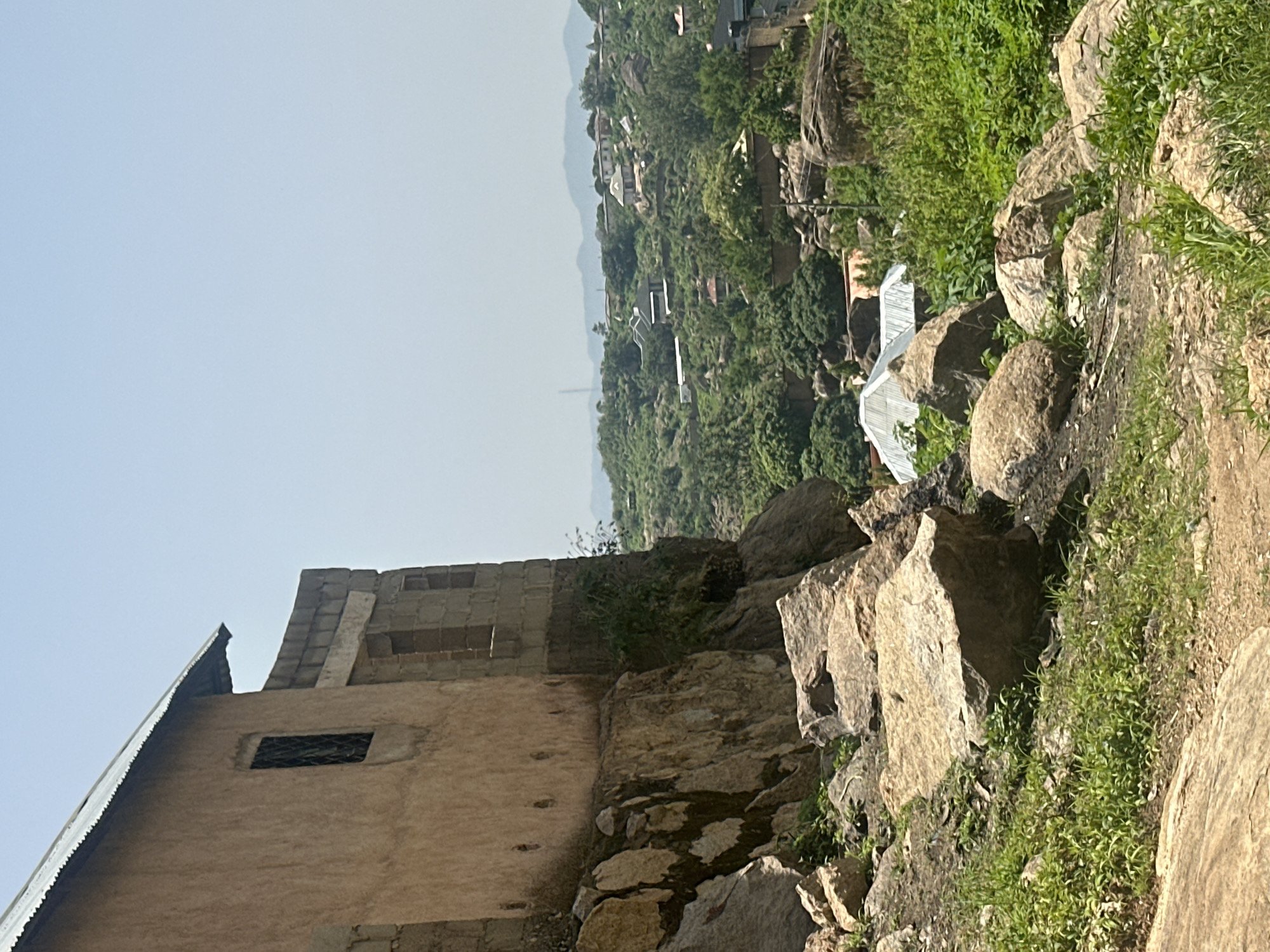 The image depicts a rugged, hilly landscape in Jos, Nigeria, under a clear, hazy sky suggesting a mid-morning or afternoon. In the foreground, a partially constructed building with a corrugated metal roof dominates the left side. The building is made of rough stone and has a few windows, one of which is visible with a dark, mesh-like covering. The wall appears to be made of a reddish-brown plaster. Adjacent to the building, large, rough-hewn rocks are scattered, covered in sparse green vegetation.
Beyond the immediate foreground, the scene opens up to a valley filled with dense green trees and more scattered rocks. Several houses are nestled amongst the trees, with some featuring white or light-colored roofs, suggesting a village or settlement built into the hills. In the mid-ground, a white, tent-like structure or greenhouse is visible among the trees and rocks, partially obscured by foliage. In the far distance, the landscape softens into rolling hills that fade into the hazy sky.
The overall impression is one of a rural or semi-rural setting in a geologically active area, characterized by rocky terrain and abundant greenery. There are no people visible in the image. The scene is peaceful and still, with no indication of any specific activity or event. The natural light suggests a warm, possibly dry day. No text is visible in the image.