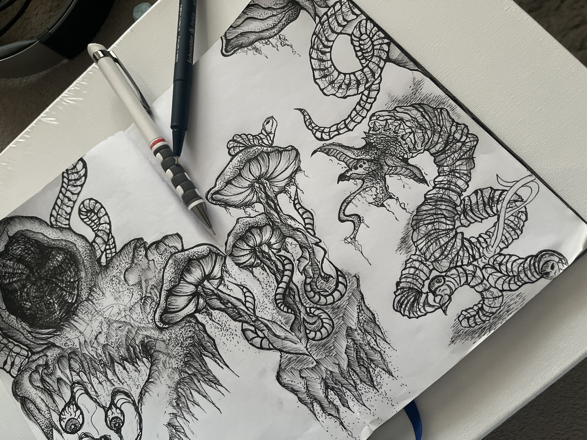 This image, captured in Sofia, Bulgaria, displays an open sketchbook featuring detailed black ink drawings of surreal and fantastical entities. Resting on the pages are two writing instruments: a white mechanical pencil or pen with a black and red striped grip, and a black fine-liner pen. Visible text on the fine-liner identifies it as "Schneider Picus" and states "Made in Germany."
The intricate artwork utilizes stippling, cross-hatching, and fine lines to create texture and depth. On the left, a large, monstrous form with a dark, cavernous eye socket, sharp, tooth-like protrusions, and multiple dangling, tear-shaped droplets each containing an eye, dominates the page. In the center, several mushroom-like structures with textured caps are depicted, intertwined with segmented, worm-like or tentacle-like appendages. To the upper right, a complex, segmented, serpentine creature with elaborate patterns coils across the page. Adjacent to it, another creature features a spiky, detailed head and a body composed of tightly wrapped, textured segments.
The scene captures a paused moment in the artistic process. The monochromatic palette highlights the detailed and often grotesque or eerie nature of the imaginary subjects, evoking a sense of dark fantasy or surrealism. The background includes a white, textured surface, possibly another art canvas or pad, and a dark, indistinct area in the upper left.