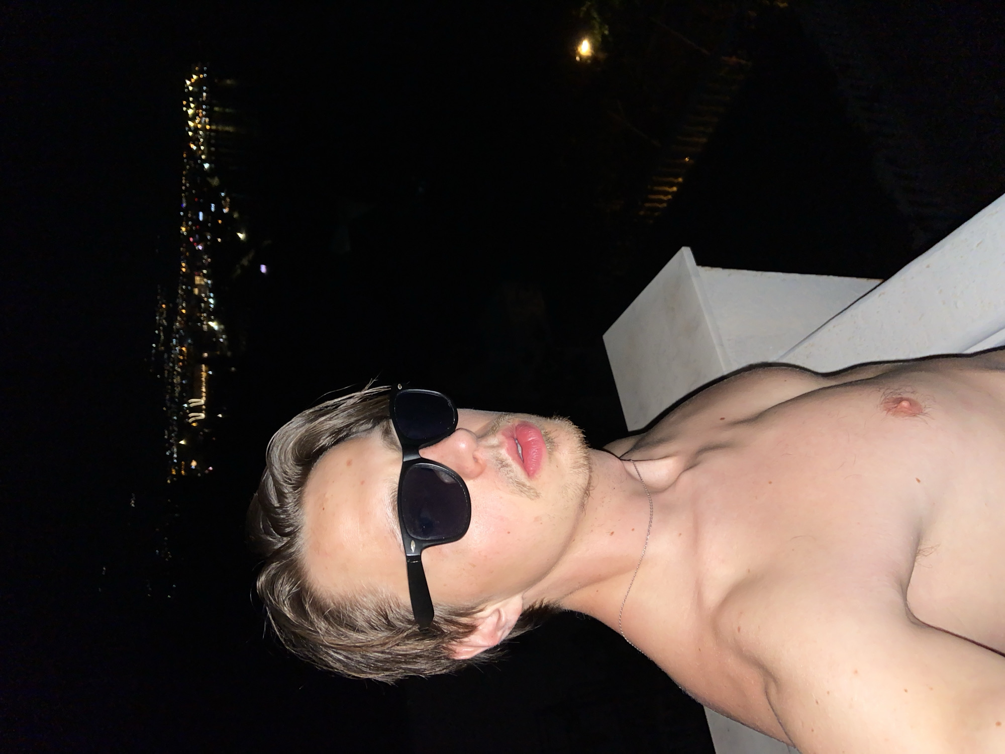 A shirtless man with sunglasses lies on a ledge at night. The city lights of Šušanj, Montenegro, are visible in the distance. The man is fair-skinned with blond hair and a light beard. He is wearing a thin silver chain around his neck. He is lying on a white concrete ledge. The background is dark.