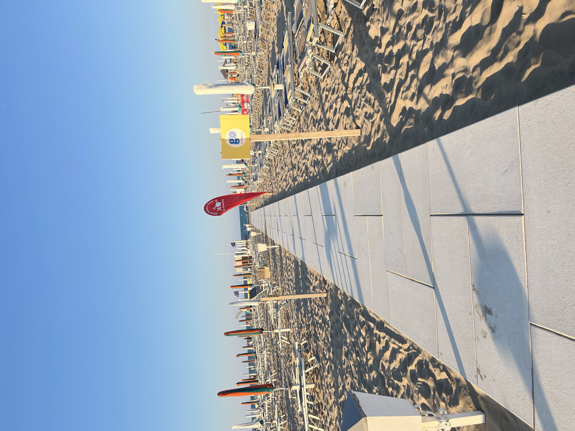 This is a ground-level, eye-level, wide shot of a paved walkway leading towards the sea on a beach in Rimini, Italy. The walkway is made of large, light-colored rectangular tiles. On either side of the walkway, rows of empty lounge chairs and closed beach umbrellas are neatly arranged on the sand. The umbrellas are a mix of colors, including orange, green, and yellow. In the mid-ground, a red flag with white text is planted in the sand, and to its right, a yellow sign with a "go" logo and text in Italian is mounted on a wooden post. The sky is clear and blue, with a few distant, faint white clouds. The sun is casting long shadows from the walkway and other objects onto the sand, indicating it is either early morning or late afternoon. There are no people visible in the image. The beach appears to be maintained and prepared for visitors.