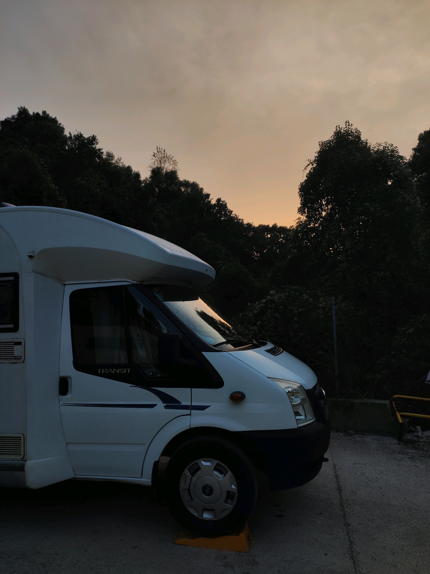 An exterior side profile shot of a white recreational vehicle (RV) parked on a paved surface during dusk. The RV, a Ford Transit, is white with dark blue trim and a dark blue stripe with a stylized design on its side. The front of the RV is visible on the right side of the frame, showing its headlights and grille. The RV is angled slightly towards the right. In the background, a dense line of dark green trees and foliage stretches across the horizon, silhouetted against a pale orange and grey sky. The sky suggests either sunrise or sunset. The ground is a rough, grey paved surface, and a yellow wedge is visible under the front wheel of the RV. A small, light-colored structure is visible on the right side of the RV, possibly part of a fence or barrier. The scene appears to be an outdoor location, potentially a campsite or parking area adjacent to a wooded area. The photograph was taken in Suera, Spain.