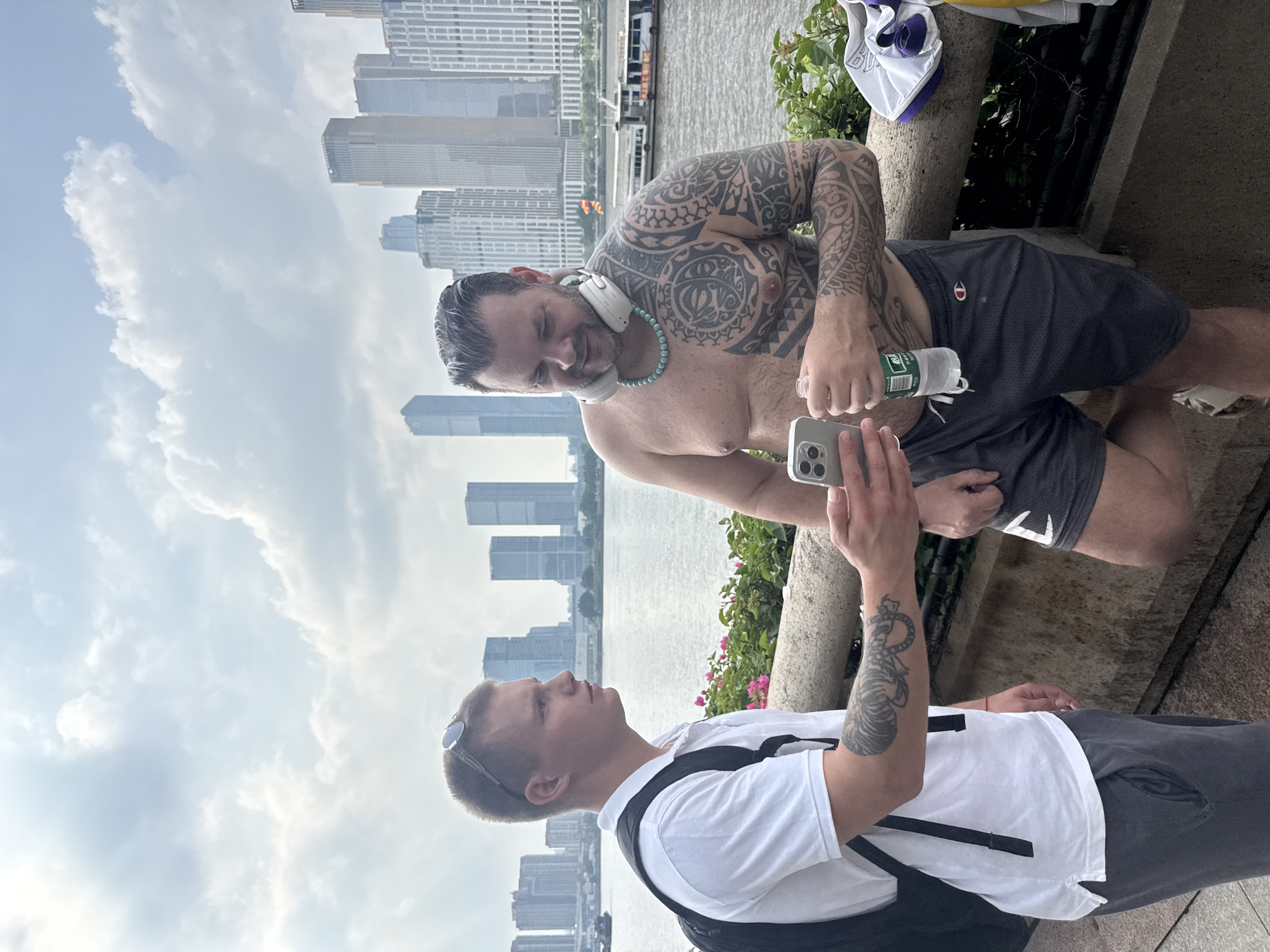 Two men are standing on a walkway next to a river in the Haizhu District of China. The man on the left is wearing a white polo shirt, dark shorts, and a black backpack. He has a tattoo on his left arm and is holding a smartphone. The man on the right is shirtless, wearing dark shorts, and has extensive tattoos on his chest and arms. He is wearing headphones around his neck and holding a bottle of water. In the background, a cityscape with numerous tall buildings and a river with boats are visible. The sky is cloudy.
