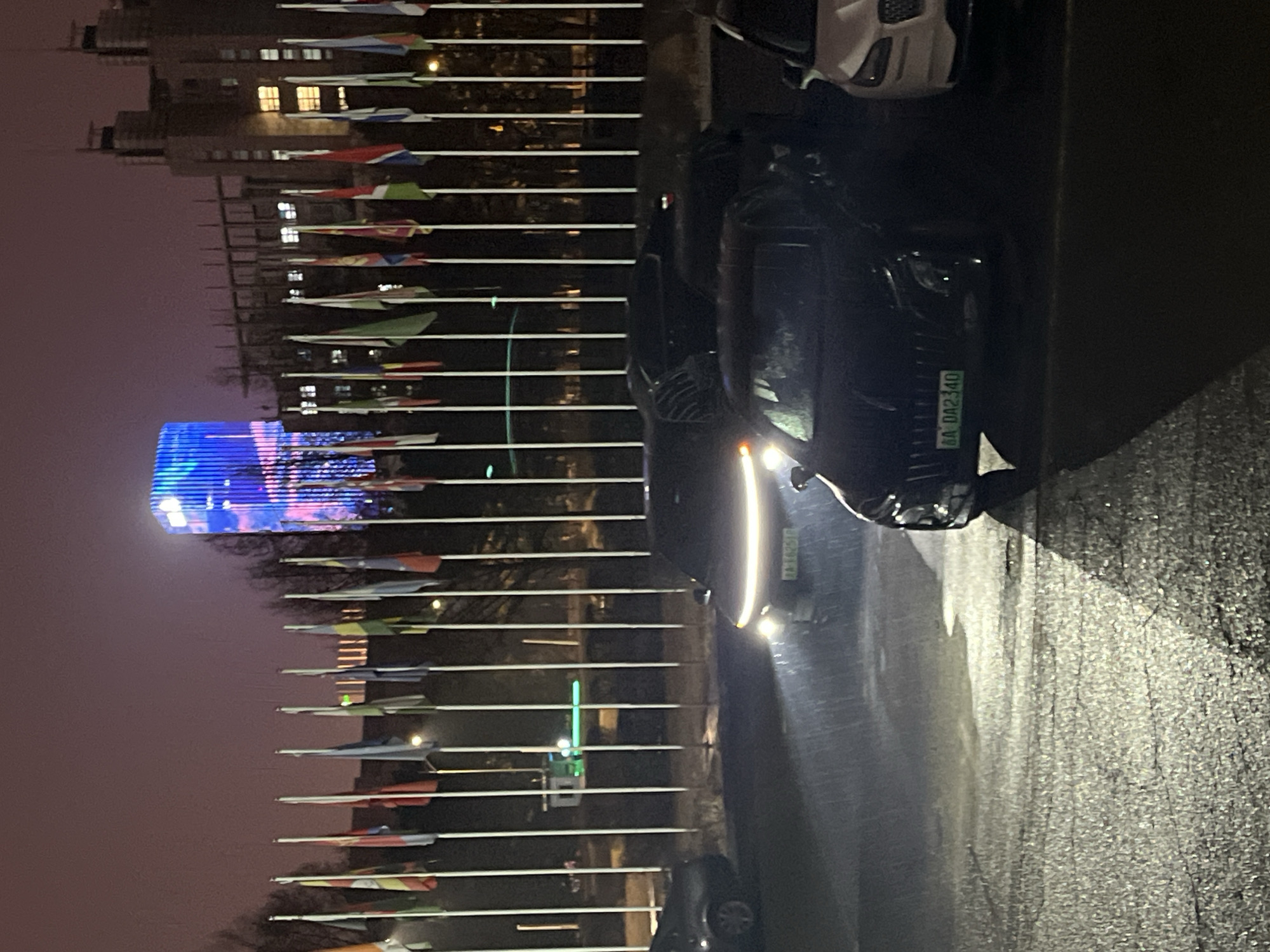 The image depicts a nighttime scene in Changchun City, China, with a line of flags and a building illuminated with blue light in the background. A dark-colored vehicle is parked on the right side of the frame. The license plate is partially visible on the car. The road surface appears wet.