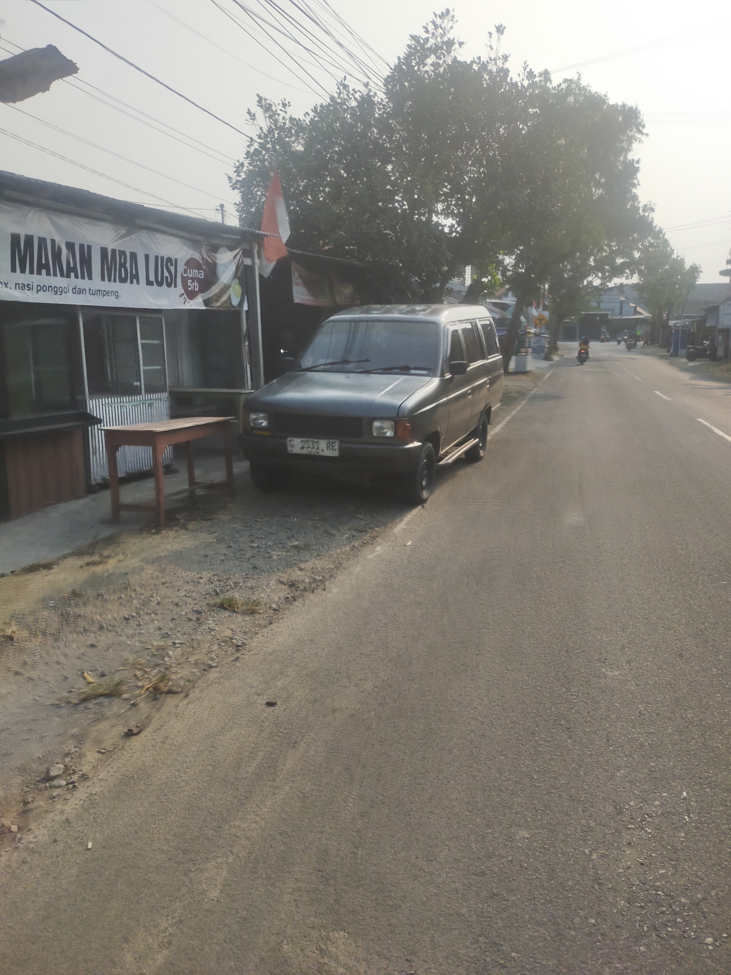 A dark green, older model SUV is parked on the side of a paved road. The SUV has a license plate that reads "G 2531 RE". To the left of the SUV is a small building with a sign that says "MAKAN MBA LUSI" and mentions "nasi ponggol dan tumpeng". There is a small wooden table in front of the building. The scene appears to be in a semi-urban or rural area of Slawi, Indonesia, as indicated by the Indonesian text on the sign and the style of buildings and road. The sky is hazy, suggesting it may be a bright or polluted day. Several motorcycles are visible on the road in the distance, indicating ongoing traffic. Trees with dense foliage line the other side of the road, and power lines stretch across the top of the frame.
