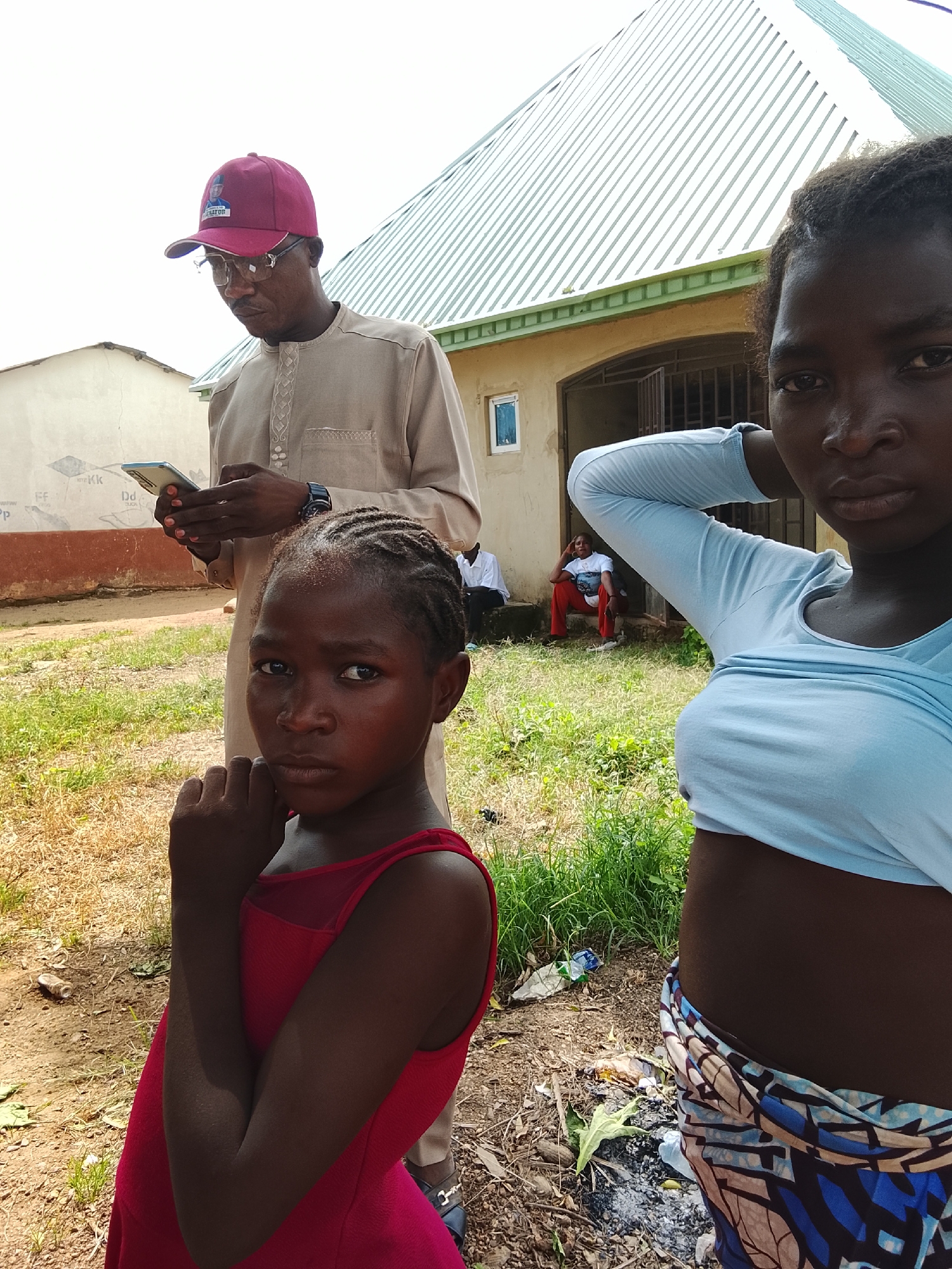 In this photograph, a group of people are gathered outdoors in what appears to be a residential area in Akwanga, Nigeria. The scene is set during the daytime, with bright sunlight casting shadows.
The primary subjects are three individuals in the foreground. On the left, a young girl with braided hair wears a bright red dress. She is looking directly at the camera with a serious expression, her right fist raised towards her chin. To her right, a man wearing a light brown traditional outfit and a maroon baseball cap with an emblem is engrossed in his smartphone. He is also wearing glasses. On the far right, a young woman is partially visible, with her arm raised behind her head and her body turned away from the camera. She is wearing a light blue t-shirt and a patterned fabric around her waist.
In the background, a building with a light-colored facade and a corrugated metal roof is visible. Two other individuals are seated on the steps in front of the building, one woman wearing a white t-shirt and red trousers, and another person whose clothing is less distinct. The ground is covered with dry grass and some scattered debris, suggesting a rural or semi-urban environment. The overall mood seems casual, though the direct gaze of the young girl and the focused attention of the man on his phone create a sense of individual introspection amidst the gathering.
There is some text visible on the baseball cap worn by the man, featuring a portrait and the word "SENATOR". Additionally, faint text and symbols are visible on the wall of the building to the left of the frame, though they are too indistinct to be read clearly.