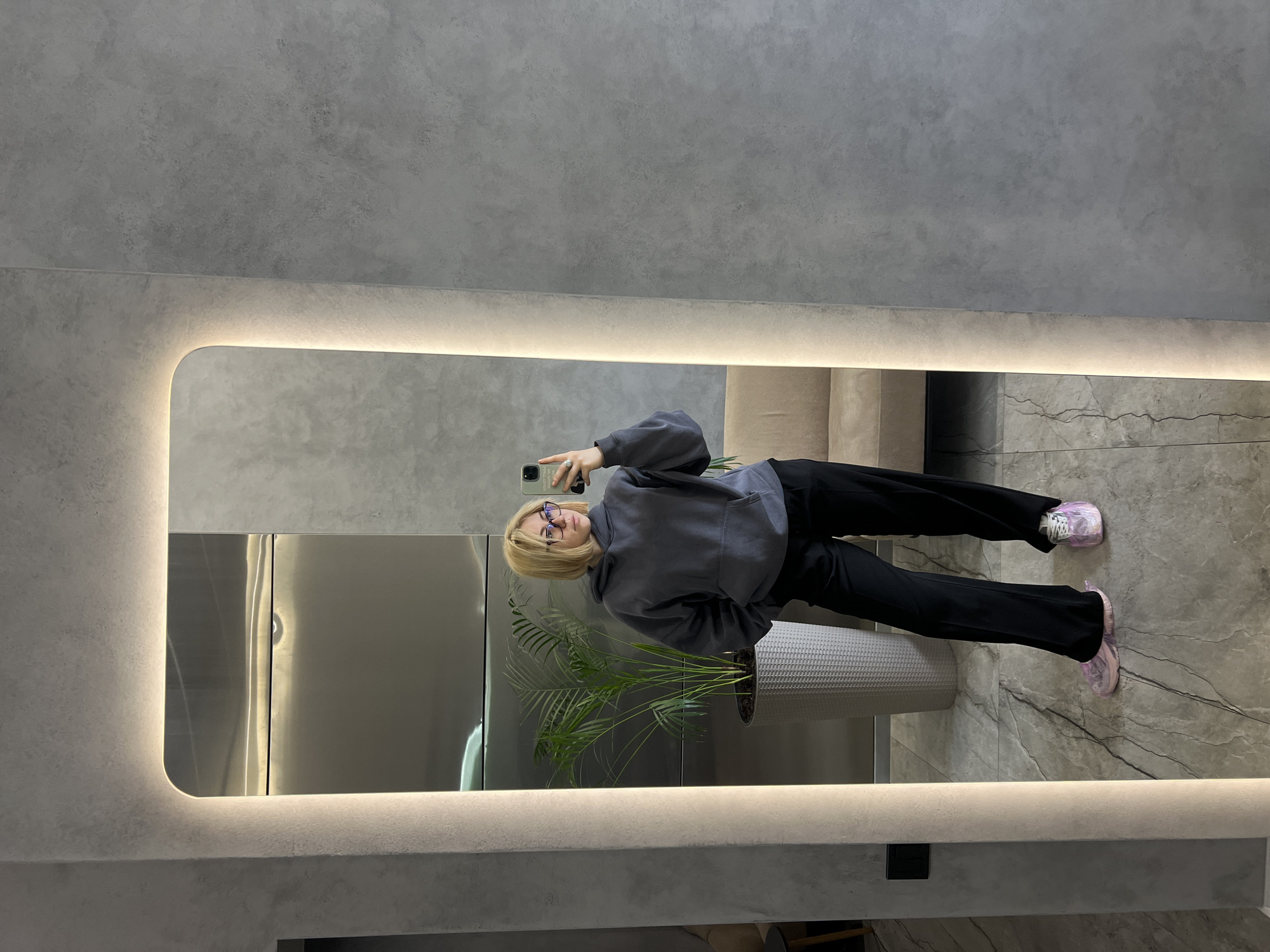 A woman is taking a selfie in a mirror. She is wearing glasses, a gray hoodie, black pants, and pink shoes with shoe covers. A potted plant sits near the mirror. The background consists of a gray wall and floor, with a glimpse of a light-colored couch. The mirror has a lighted frame. The location is likely a private residence or interior space in Odesa, Ukraine, given the query's contextual information.
