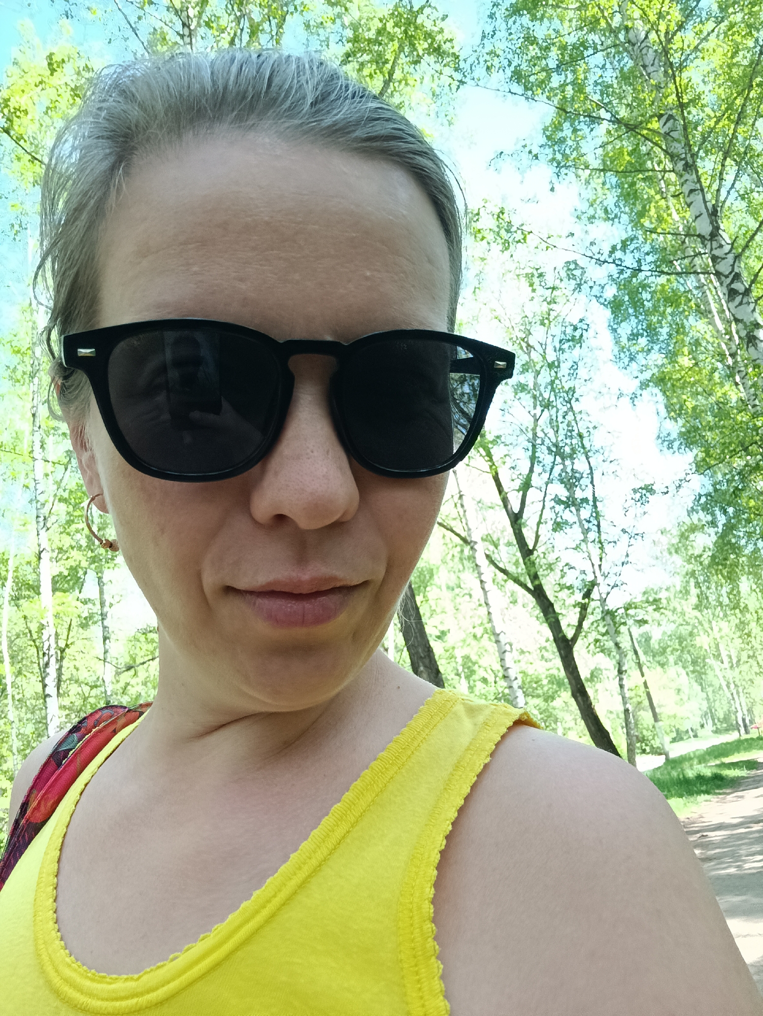 A woman with short, gray hair, wearing black sunglasses and a yellow tank top, poses for a selfie in a park. The background shows birch trees with green leaves, suggesting a park in the spring or summer. The trees appear to be located in a park in the city of Chaikovsky, Russia.