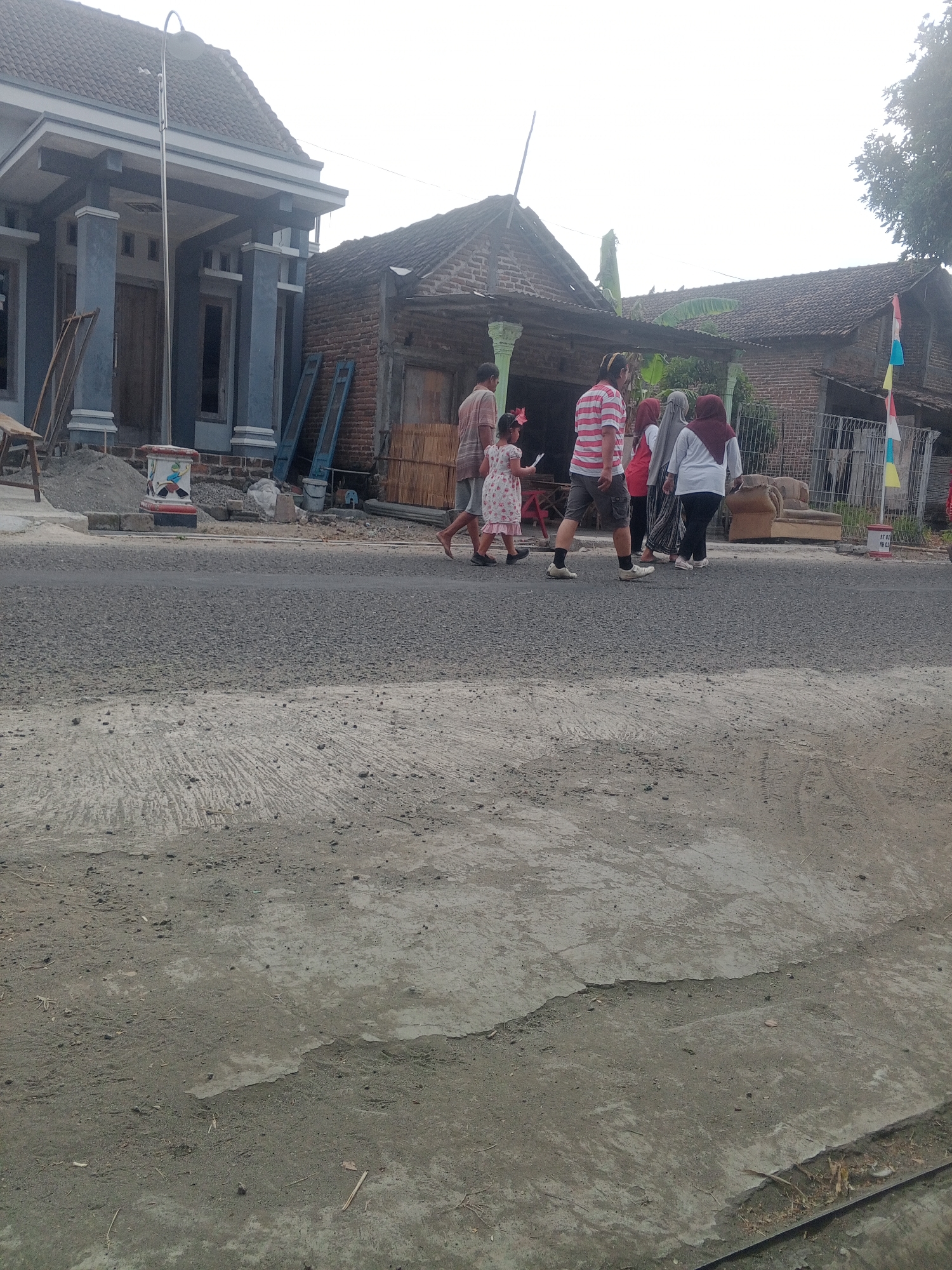 In Lembah, Indonesia, a group of six people are walking across a paved road, with buildings and construction visible in the background. The group consists of three adults and three children. One adult is wearing a red and white striped shirt, and another is wearing a gray hijab. A young child, possibly a girl, is wearing a patterned dress. They are walking from left to right across the frame.
Behind the group, there are several buildings, including a partially constructed house with blue pillars and a gray roof. Adjacent to this is a smaller, older building with a corrugated iron roof and brick walls. Construction materials, such as piles of sand and wooden scaffolding, are scattered around the area. To the right of the buildings, a faded brown sofa is visible. The road surface is asphalt, with some patches of concrete or dirt on the side. The sky is overcast, and the overall lighting suggests it is daytime, possibly morning or late afternoon. The scene depicts a common street view in a residential area undergoing some development.