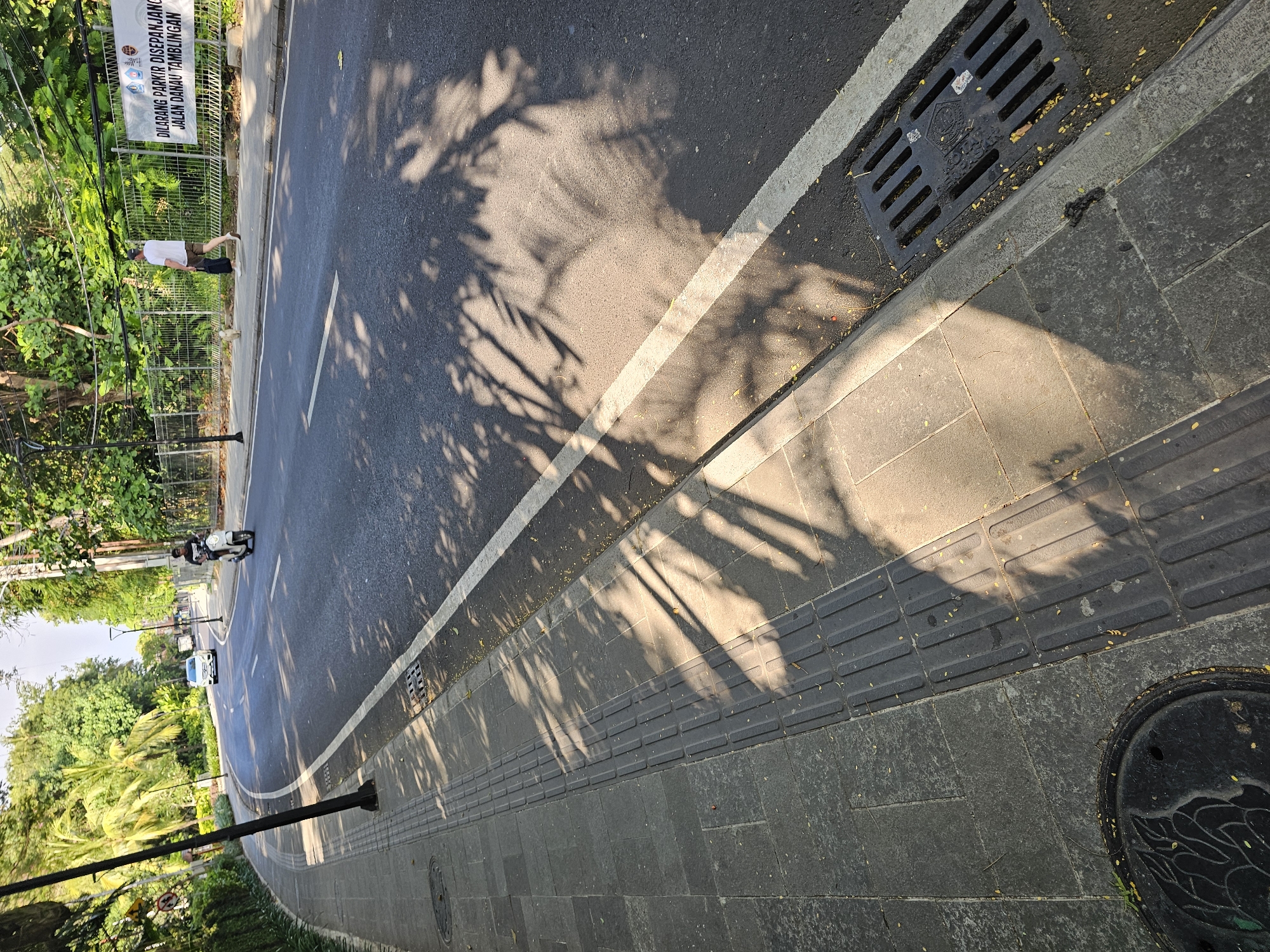 The image shows a sunlit street scene in Denpasar, Indonesia. A paved road with a white lane marker is visible, partially shaded by tree shadows. A sidewalk borders the road, featuring tactile paving designed for visually impaired pedestrians. A man in a white shirt and dark pants stands near a sign prohibiting parking on Jalan Danau Tamblingan. Further down the road, a scooter and car are present. A storm drain is built into the curb. A manhole cover is set into the sidewalk.