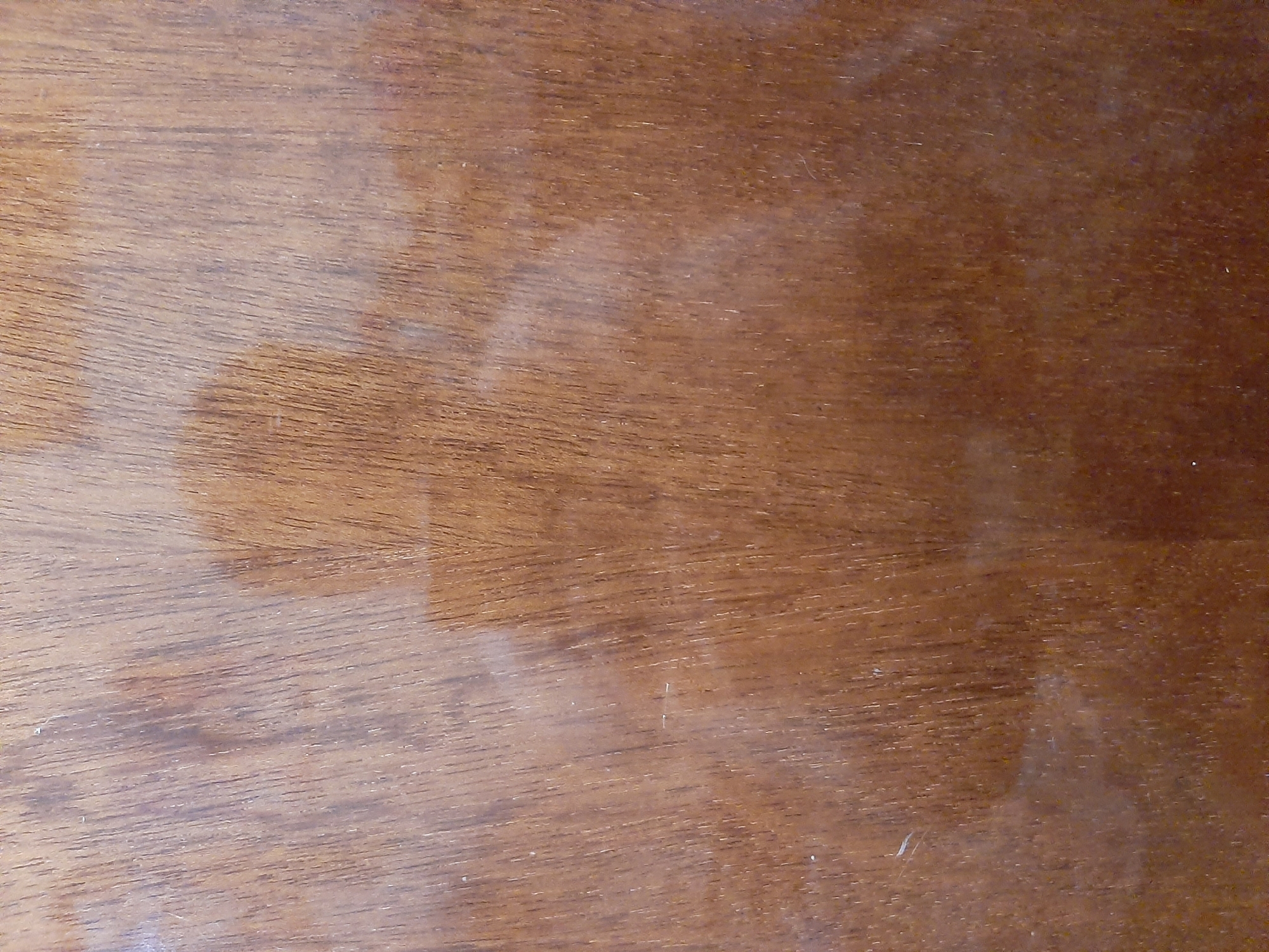 The image is a close-up, vertical, eye-level shot of a wooden surface. The wood grain is predominantly vertical with some slight diagonal variations. The color is a rich, medium brown with darker streaks and lighter, almost golden highlights scattered throughout. The surface appears to be finished, with a slight sheen that reflects light, creating subtle bright spots and a hazy, diffuse reflection in the center of the frame. This reflection is faint and indistinct, suggesting a person or object was present but is not clearly defined. The texture of the wood is visible, showing fine parallel lines and occasional subtle imperfections. There are a few very small, light-colored specks on the surface, which could be dust or minor blemishes. The lighting is soft and diffused, casting no harsh shadows.