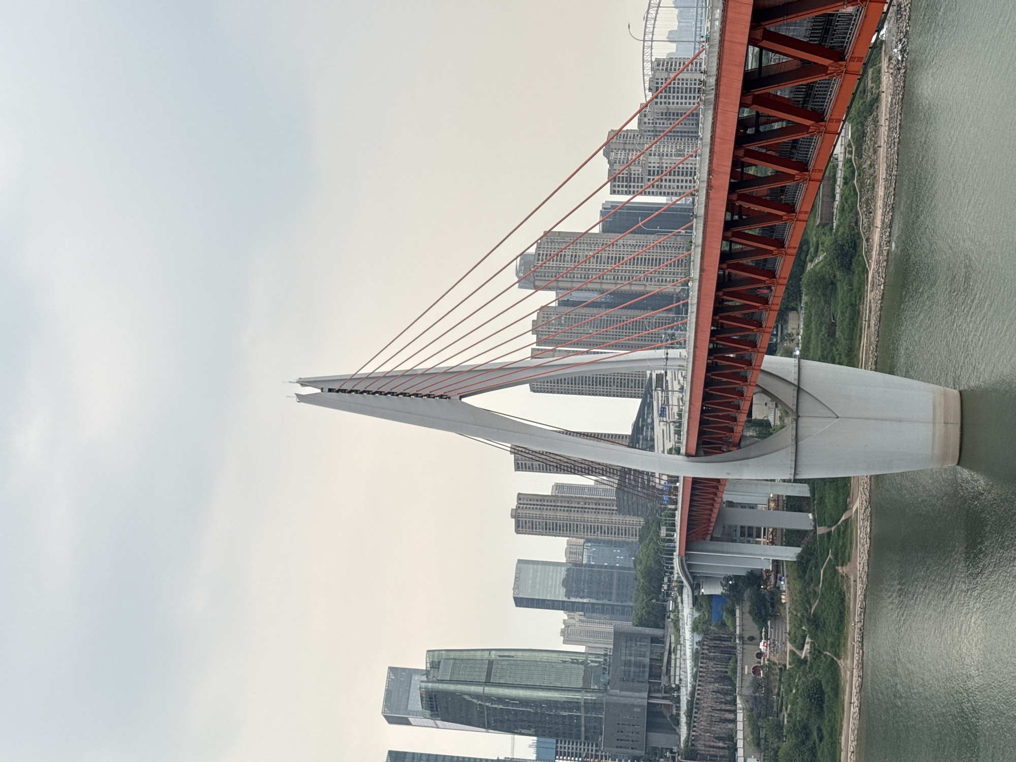 The image captures a segment of the iconic Chongqing's Dongshuimen Bridge, a distinctive red cable-stayed bridge in Yuzhong District, China. The bridge is predominantly red with white support structures. Visible are the steel cables anchoring into the bridge's support structure. In the background, tall skyscrapers can be seen. The bridge spans over water, with a park-like green area visible along the bank. The sky is overcast, suggesting potentially hazy conditions.