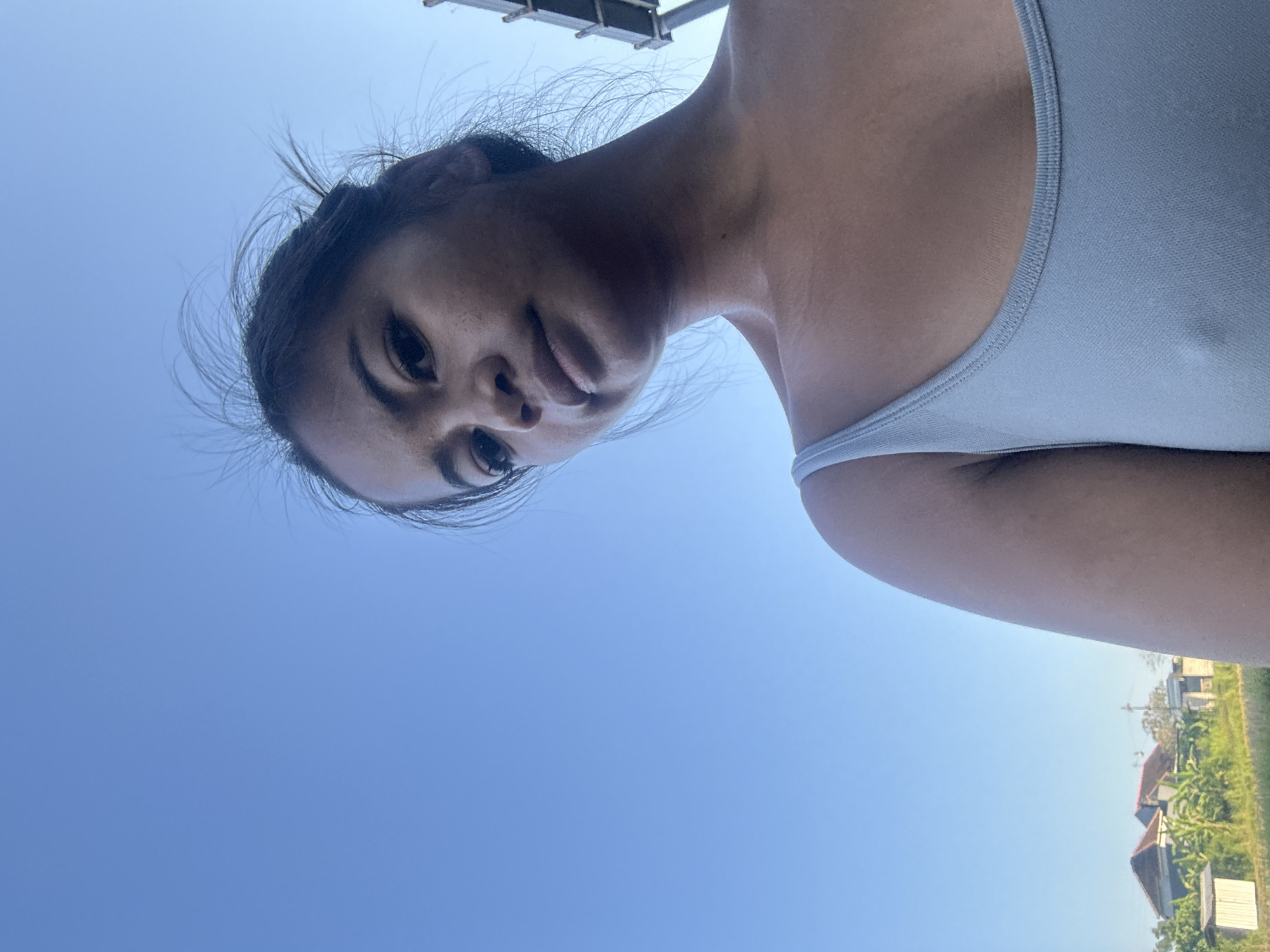 A low-angle, outdoor, medium shot features a young woman with dark hair and brown skin wearing a light blue tank top against a clear, bright blue sky. She is looking slightly down and to her right, with her hair loosely tied back and wisps of hair floating in the breeze. Her face is in sharp focus, with a hint of a smile. In the background, to the left, there is a rural scene with houses, trees, and fields under the same blue sky. The overall impression is a candid moment captured in a natural, outdoor setting, possibly in the city of Sukawati, Indonesia, given the tropical vegetation and style of the houses.