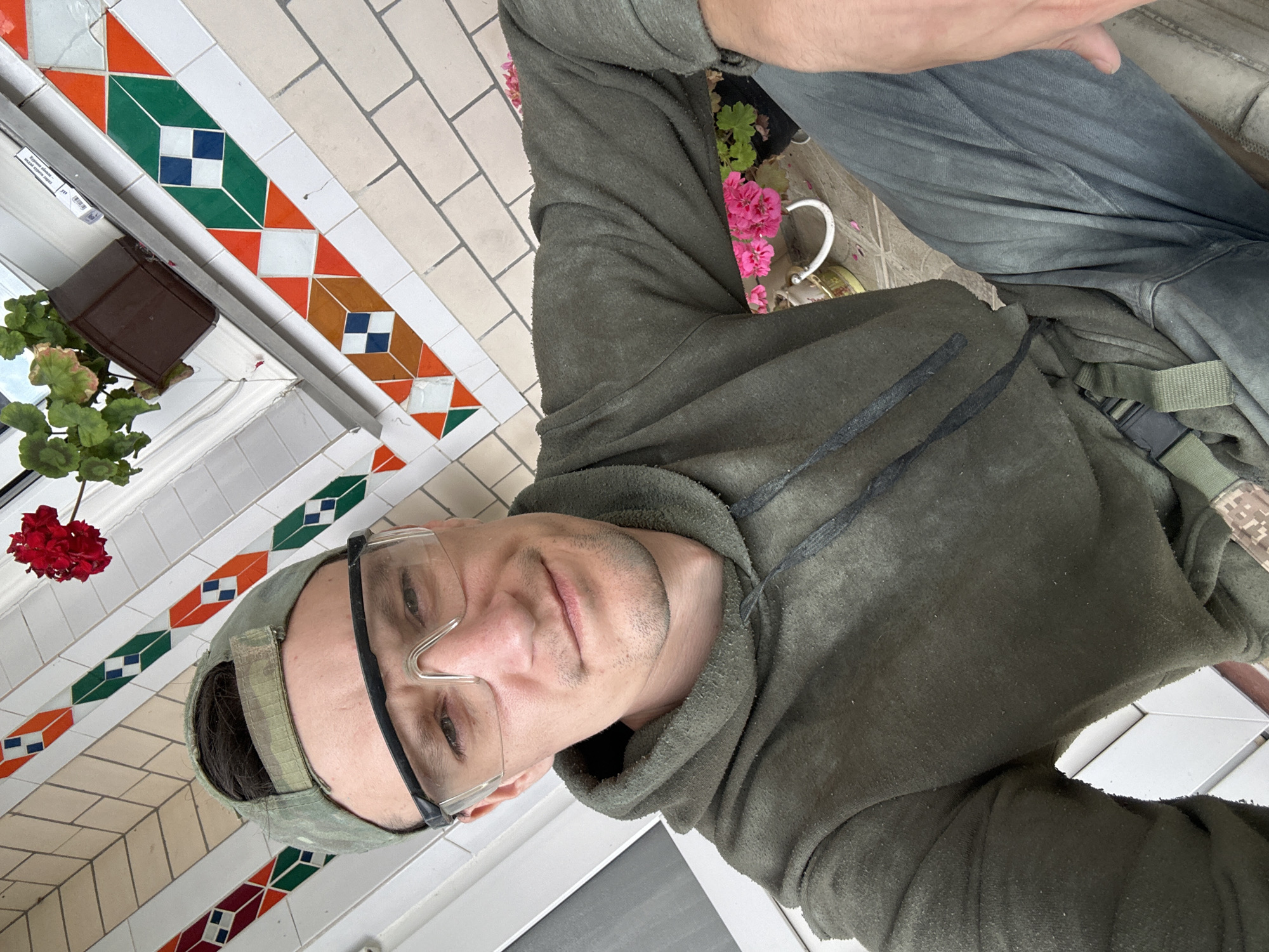 A man wearing a camouflage baseball cap, clear protective goggles, a dark olive green hoodie, and dark pants is sitting down outdoors. He is looking directly at the camera and smiling slightly. Behind him, there is a tiled wall with an ornate pattern of orange, green, and white geometric shapes. A potted red flower is visible to his right, and a plant with pink flowers is also present in the background. The man's right hand rests on his leg. The scene suggests a casual, possibly DIY or gardening context, and the tiled wall could be part of a house or building exterior. The location is identified as Makiv, Ukraine.