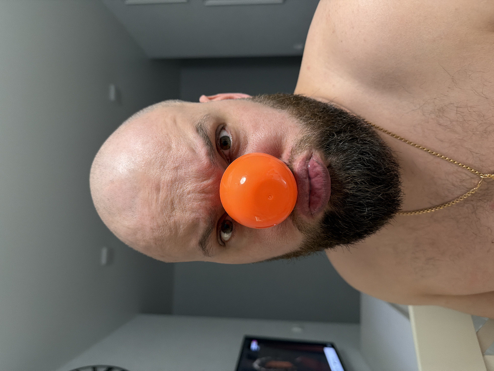 A close-up, rotated portrait shows a bald man with a beard wearing an orange plastic ball on his nose. He has light skin, dark eyes, and a gold chain around his neck. The background is a gray wall and a partially visible television screen. The image does not contain any information about the city of Votkinsk, Russia.