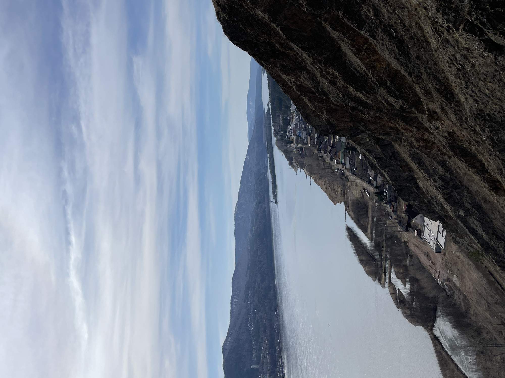 The image shows a view of the Yenisey River and the city of Krasnoyarsk, Russia, from the top of a rocky cliff. The river flows in the center of the frame, with a line of buildings along the right bank. A mountain range is on the left side of the river. The sky is light blue with thin white clouds. A dark, craggy rock formation is in the foreground on the right, partially obscuring the view.
