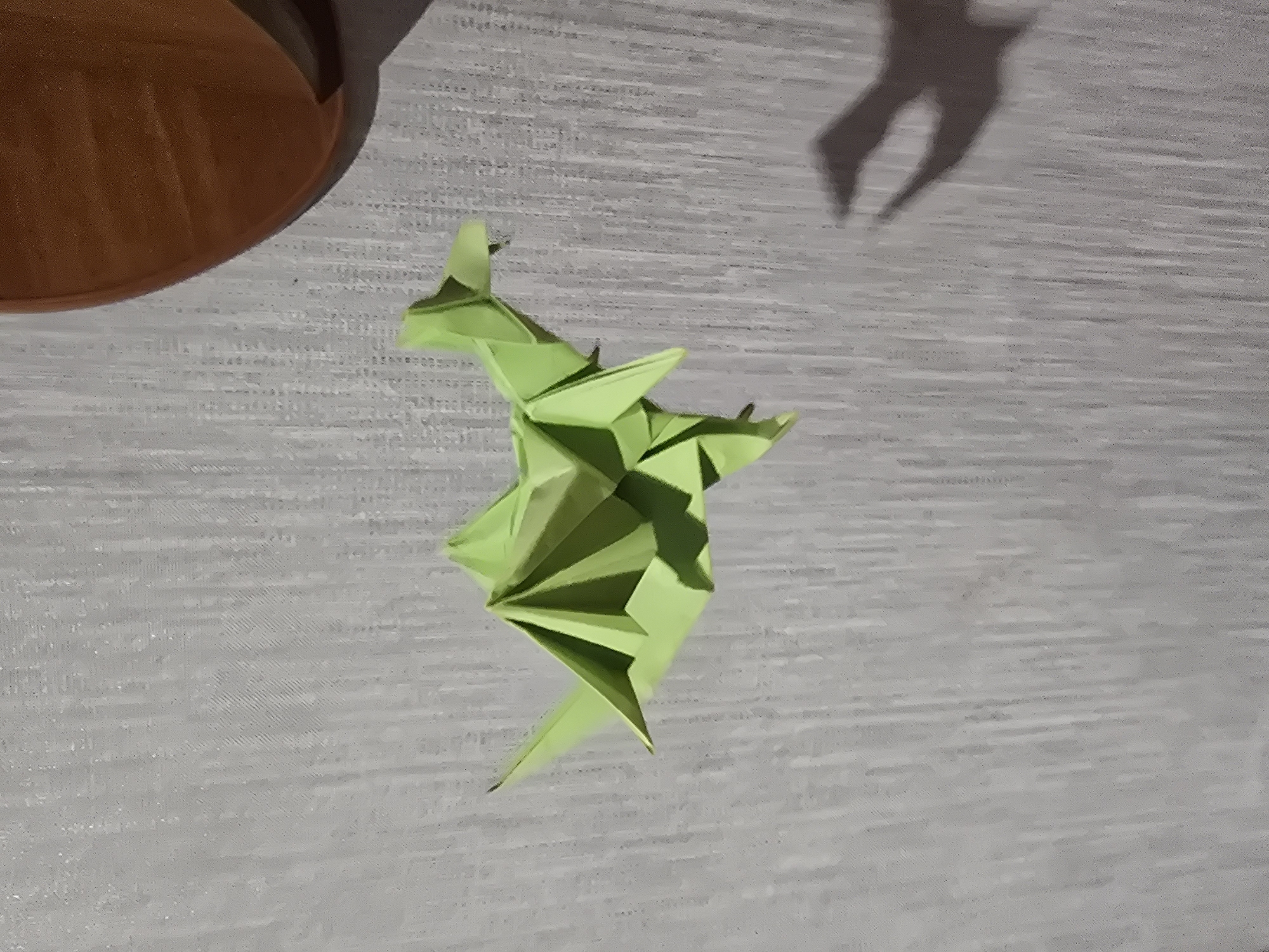 This is an overhead, medium-shot, indoor photograph of a green origami dragon sitting on a textured grey surface. To the left of the origami dragon is the side of a brown, bowl-shaped object, possibly a pot. To the upper right of the dragon, a dark grey shadow of what appears to be the same origami dragon is cast on the grey surface. The light source appears to be to the left and above the scene, casting the shadow of the dragon and a portion of the brown object. The overall lighting is dim. The composition is slightly tilted, with the dragon positioned in the center of the frame, angled towards the bottom right. No text is visible in the image.