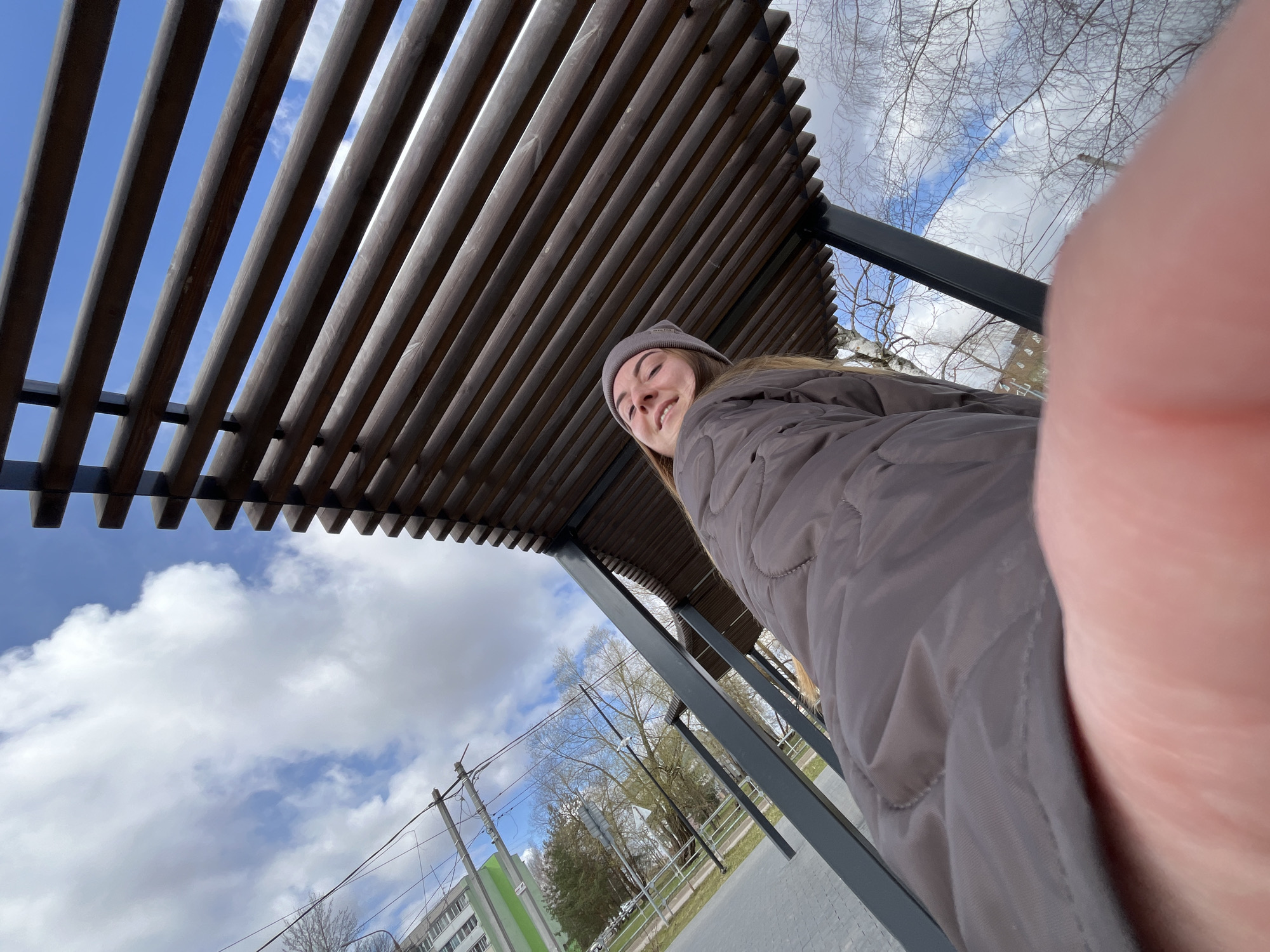 A low-angle shot features a woman in a grey beanie and a grey quilted jacket, smiling towards the camera. A finger partially obscures the right side of the frame. Above her is a pergola made of wooden slats. The background includes a blue sky with white clouds, trees, power lines, and a building. The location is likely in Vysokovsk, Russia, as per the metadata.