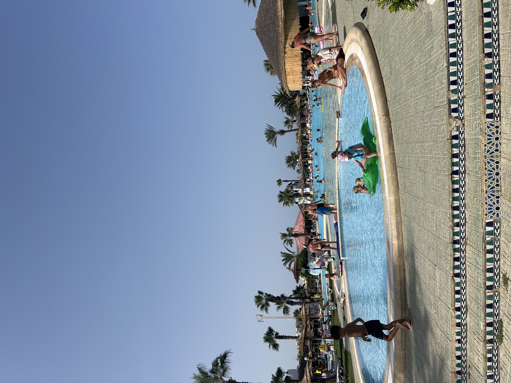 The image depicts a lively scene at a resort swimming pool in Hiboun, Tunisia. The bright blue water of the pool is filled with numerous people of various ages enjoying the sunny day. Several individuals are swimming or wading, while others are relaxing on inflatable toys, including a green alligator. A young boy in dark swim trunks walks along the pool's edge in the foreground. Surrounding the pool are lounge chairs and umbrellas, and in the background, palm trees and resort buildings are visible. The sky is clear and blue, indicating pleasant weather.
