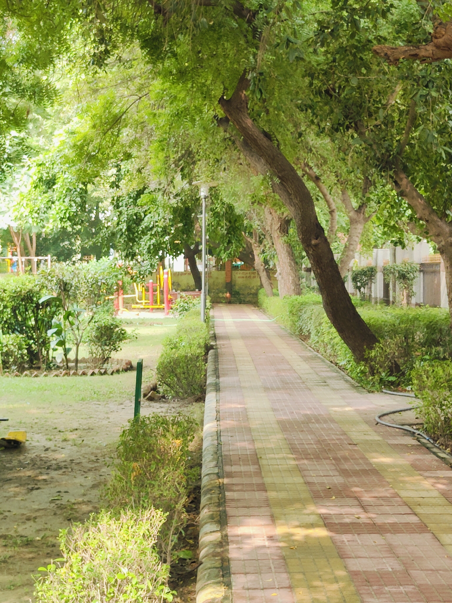 The image depicts a paved pathway in a park in Gurgaon, India. The pathway, made of light and dark pinkish-tan tiles, runs diagonally from the foreground to the background, lined by lush green bushes on the right. Sunlight dapples the pathway, suggesting it is daytime. On the left side of the pathway, there is a grassy area with several small trees and some playground equipment in the middle ground, primarily red and yellow. Tall trees with dense green foliage arch over the pathway, creating a shaded canopy. A lamppost is visible on the right side of the pathway. The overall environment appears to be a well-maintained public park, likely a recreational area within a residential or urban setting in Gurgaon. No people are visible in the image.