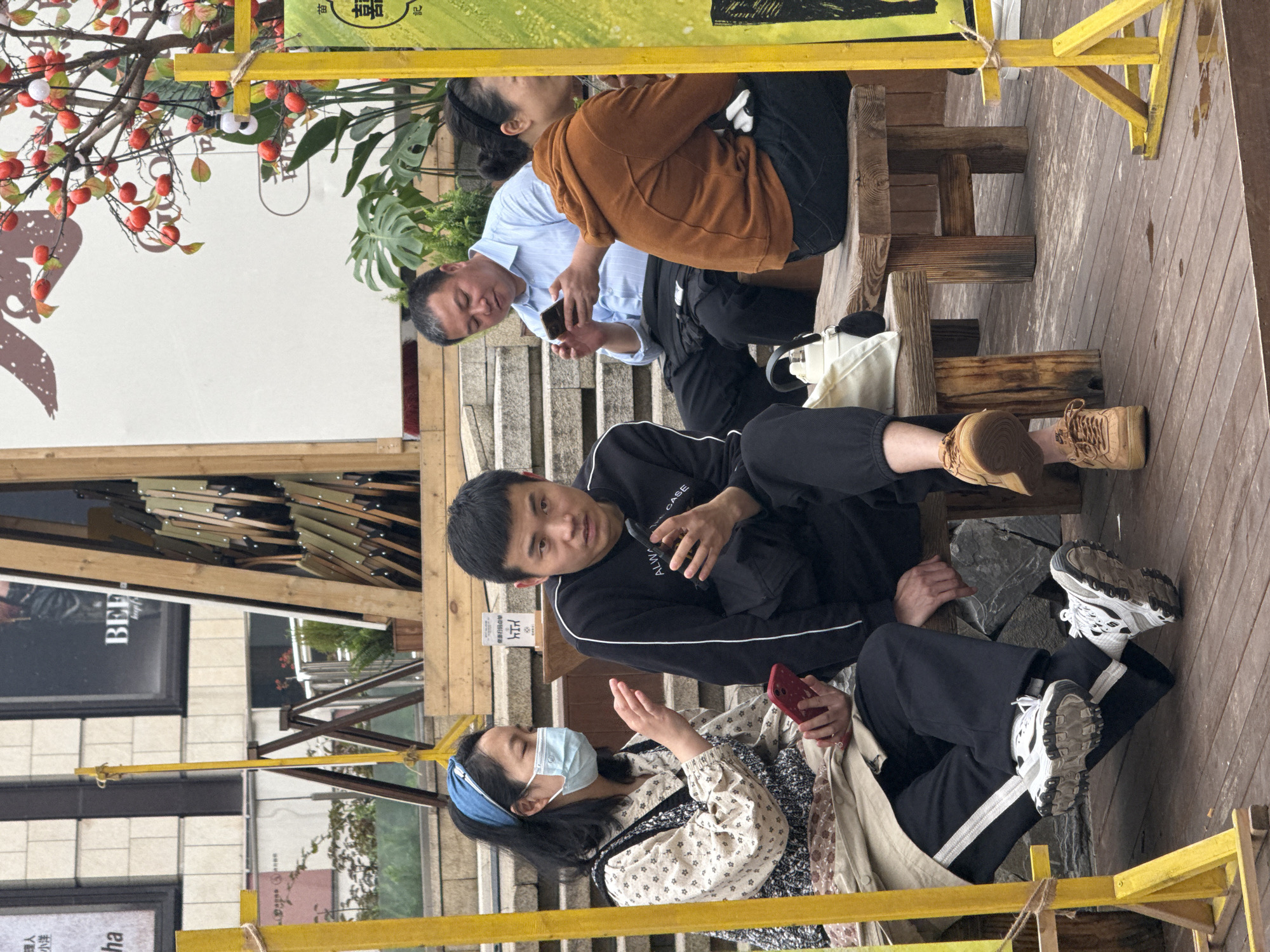 In the Yuzhong District, China, an outdoor setting depicts a group of four individuals seated at wooden benches beneath a yellow frame structure, seemingly engaging in social interaction or using electronic devices. A woman with a blue headband and a medical face mask holds a red smartphone, while a man in a black tracksuit sits beside her. Above them, a man in a light blue shirt is looking at his smartphone and conversing with a woman in an orange sweater. In the backdrop, an advertisement sign for "BEE keep" is visible, along with decorative elements such as plants, red berries, and foliage, contributing to the ambiance of the location. Wooden steps and flooring are also visible.