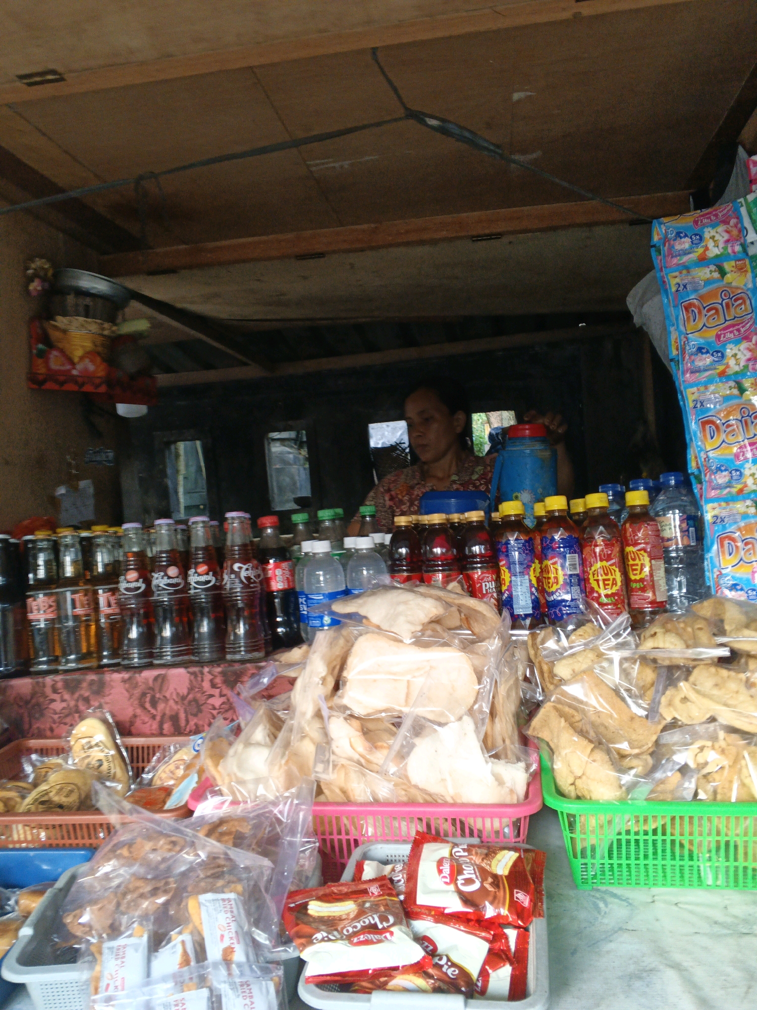 This is a medium shot, eye-level, outdoor photograph of a small street-side stall in Manggis, Indonesia. The stall appears to be made of unfinished wood with a corrugated metal roof. Inside the stall, a woman with dark hair pulled back is visible behind a display of various goods. She is wearing a patterned shirt. In front of her, on shelves and in baskets, are numerous items for sale.
The display includes:
* Several rows of bottled drinks, including what appear to be sodas and possibly teas or juices, with labels in various colors. Some bottles are red, others are clear with amber liquid.
* Bags of snacks, some appearing to be crispy crackers or chips, are displayed in plastic bags and pink and green plastic baskets.
* Packets of food items, possibly snacks or condiments, are visible in the foreground, some with red and white packaging.
* A large blue thermos is positioned behind the woman.
* A small decorative item, possibly a flower arrangement or shrine, is on the left side of the stall.
* Large plastic bags of what appear to be laundry detergent or fabric softener, with "Daia" branding and floral patterns, are hanging on the right side of the stall.
The lighting suggests it is daytime, possibly overcast or with partial shade from the stall’s roof. The ground in the foreground is a textured surface, possibly concrete or a similar material. The overall scene depicts a typical small retail operation in a Southeast Asian setting.