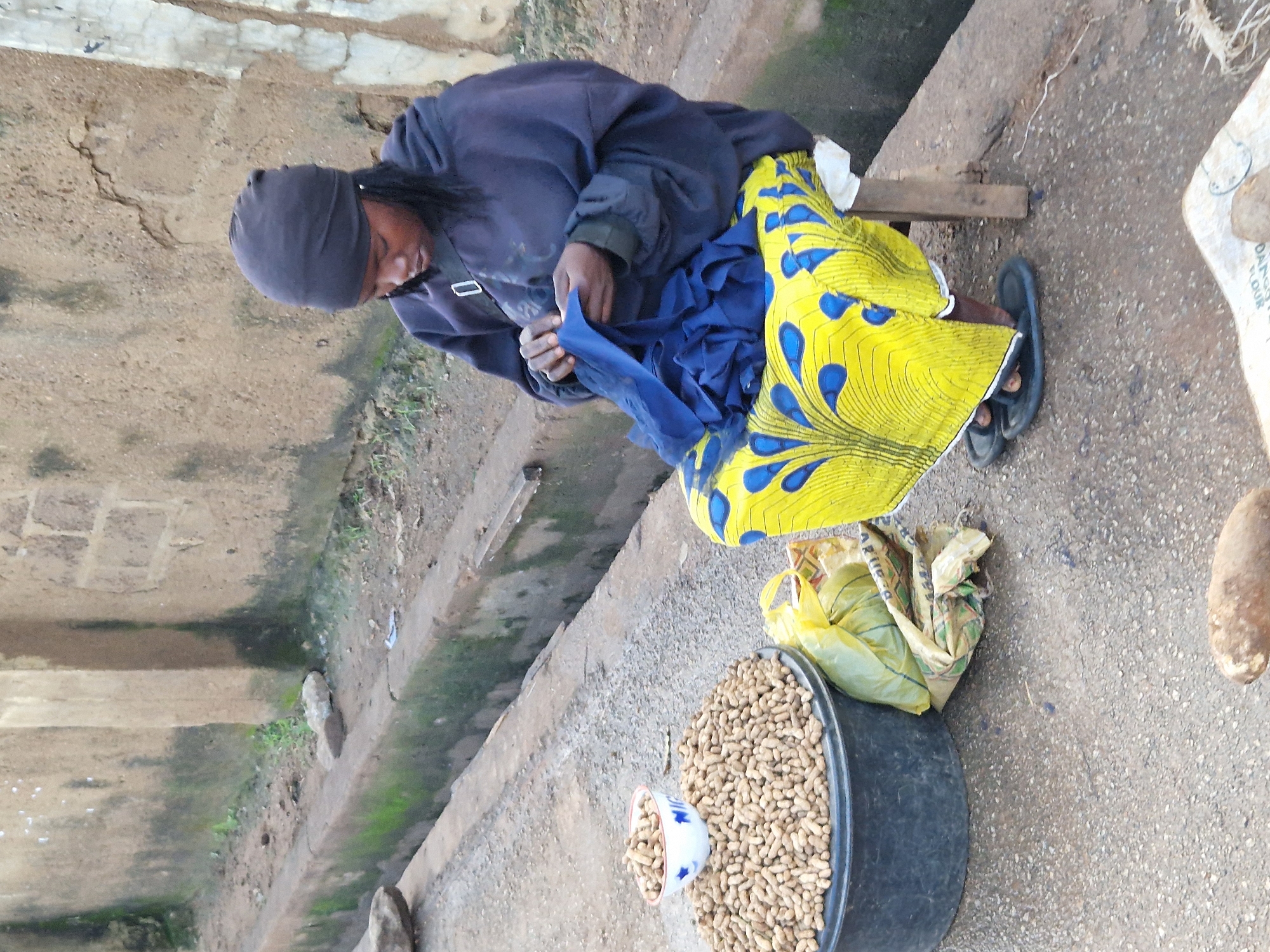 A Black woman sits on a wooden stool, hunched over as she stitches a piece of blue fabric. She wears a dark headscarf and a dark hooded jacket, and a vibrant yellow and blue patterned skirt. In front of her, a large black tub overflows with peanuts, with a small white bowl containing more peanuts resting on top. Next to the tub, a crinkled yellow bag lies on the ground. The scene is set against a rough, light-brown textured wall, possibly a building facade, with a drainage channel running along the ground in front of it. The ground surface is paved with small stones. The environment appears to be an outdoor market setting in the city of New Stadium, Nigeria.