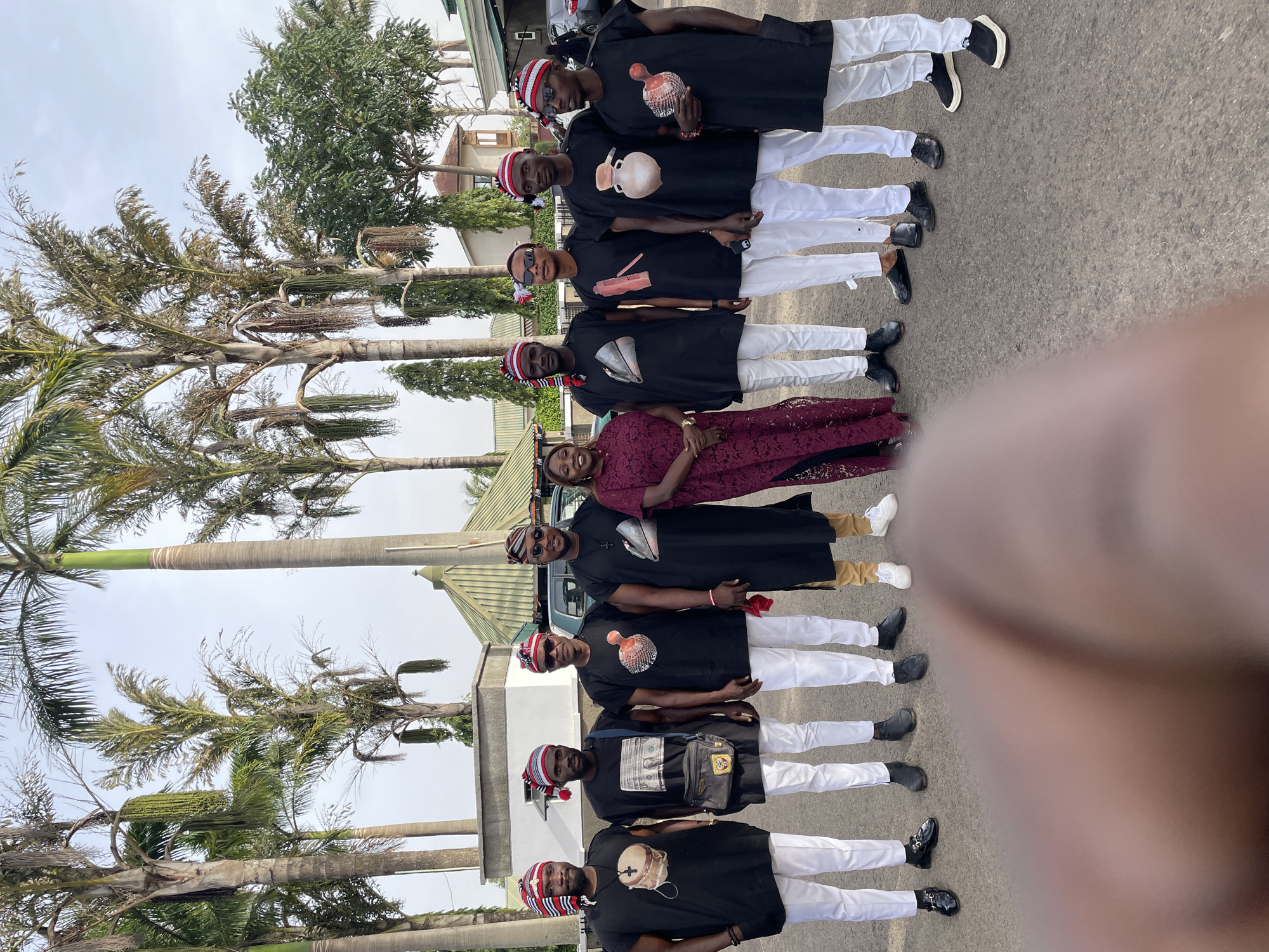 A photograph shows eight people standing outdoors. Seven men are wearing matching traditional clothing with red-and-white striped hats, black tops with various cultural images printed on the front, and white trousers. The eighth person, a woman, is standing in the middle wearing a patterned maroon dress. The group is standing on a paved surface, likely a road. Palm trees and buildings are visible in the background. The location is likely in Jos, Nigeria, due to the clothing and vegetation.