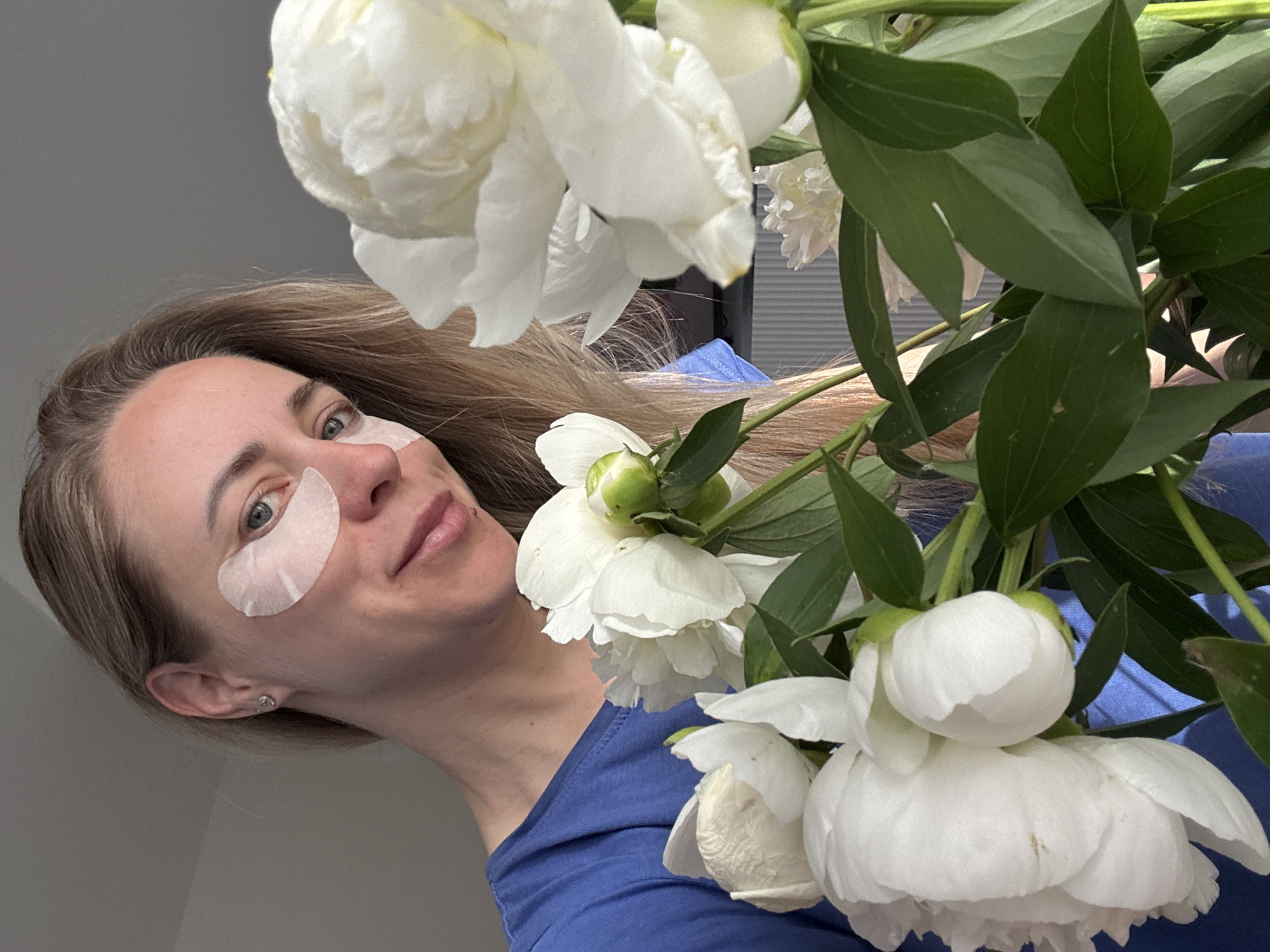 A woman with blonde hair and under-eye masks is holding a bouquet of white peonies. She is wearing a blue t-shirt and is looking at the camera with a slight smile. The background is blurred and appears to be an indoor setting. The image does not provide any information about the location in Chaikovsky, Russia.