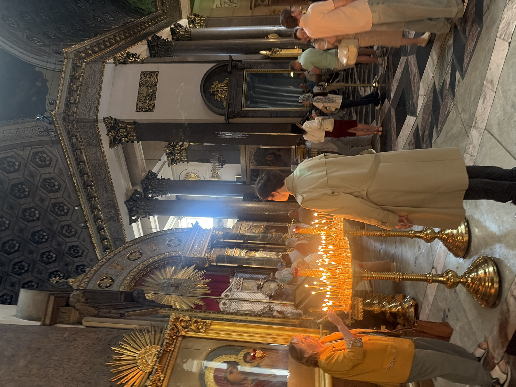 An indoor scene captures a religious interior with multiple individuals. In the foreground, a woman in an orange jacket is seen tending to a large golden candelabra filled with lit candles. To her right, another person in a tan trench coat stands facing away from the camera, also looking towards the candelabra.
The background reveals a grand architectural space with ornate columns and a decorative ceiling. Several other people are dispersed throughout the area, some appearing to be visitors or congregants. Sunlight streams in from an unseen source, casting bright light and shadows. The floor is a mosaic of geometric patterns in dark and light stones. The overall atmosphere suggests a place of worship or historical significance.