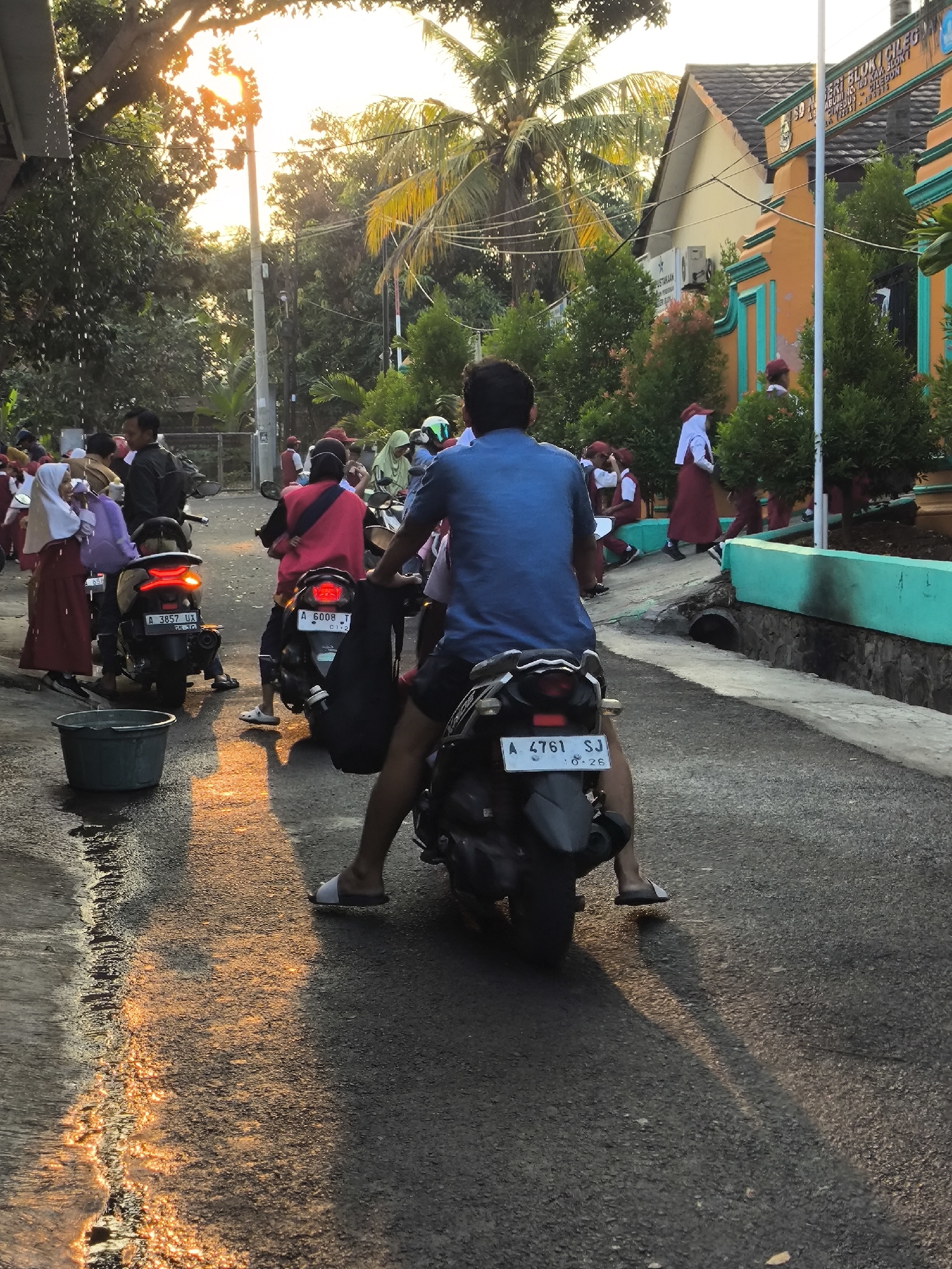 The image depicts a street scene in Cilegon, Indonesia, with a group of people and motorcycles. In the foreground, a person wearing a blue shirt is sitting on a motorcycle, facing away from the camera. The motorcycle has a license plate reading "A 4761 SJ .8.26". To the left, another motorcycle with license plate "A 3857 UX .8.16" is visible, with a person in a pink vest riding it. Several individuals, including children in school uniforms consisting of maroon skirts and white hijabs or shirts, are gathered on the side of the road. The background shows buildings with orange and green facades, palm trees, and dense foliage, suggesting a tropical environment. The setting sun casts long shadows and illuminates the wet asphalt, indicating it may have recently rained. A green plastic tub is placed on the left side of the road. The overall atmosphere appears to be one of a school or community gathering, possibly at the end of the school day.
