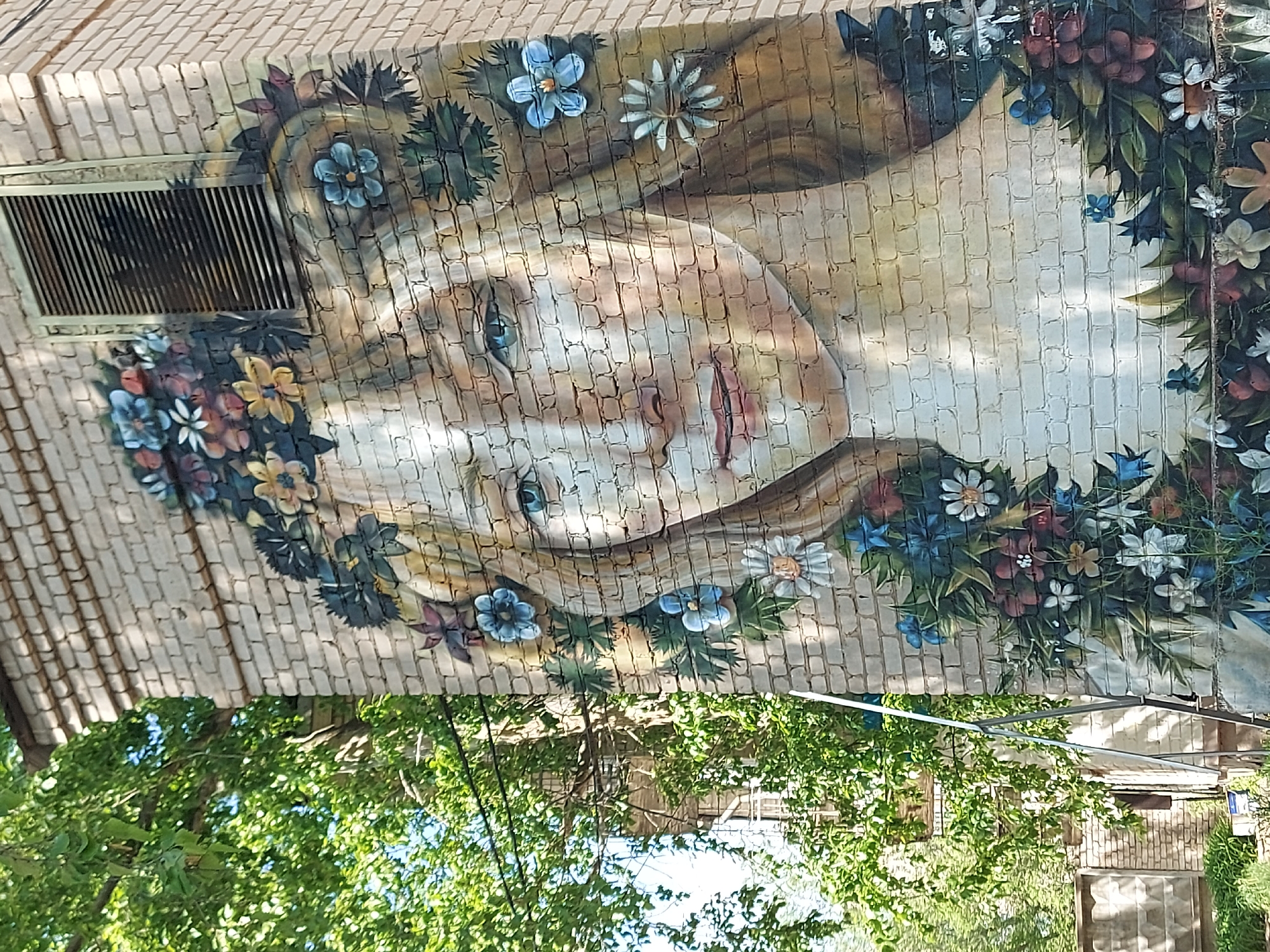 The image depicts a mural of a woman with flowers in her hair, painted on a brick wall. The mural is located on a building in Chaikovsky, Russia. The woman has fair skin, blonde hair, and blue eyes. She is surrounded by a wreath of flowers. The image is partially obscured by trees and foliage. A building with a white door is visible in the background. A metal grill is visible on the left side of the wall.