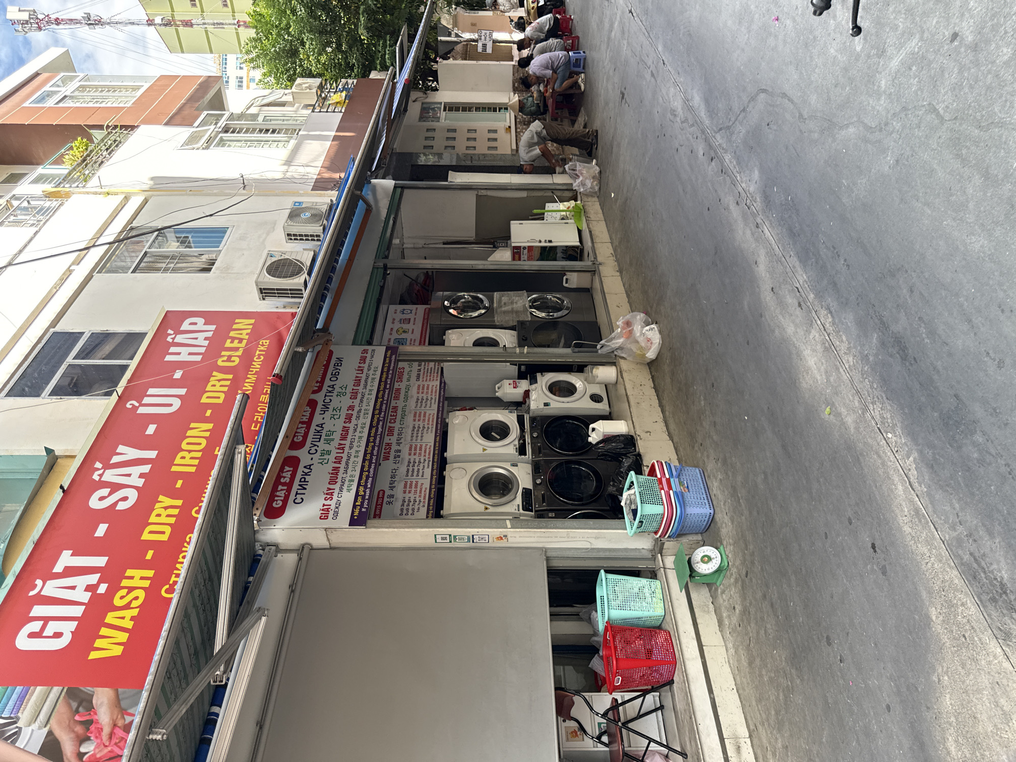 This is a medium-shot, exterior photograph of a laundromat on a street in Nha Trang, Vietnam. The laundromat's facade is bright red and yellow, with Vietnamese and English text indicating services offered, such as "GIẶT - SẤY - ỦI - HẤP" and "WASH - DRY - IRON - DRY CLEAN". Below the main sign, there is a smaller sign with pricing information in multiple languages. The storefront features several commercial-grade washing machines and dryers stacked on top of each other. In front of the laundromat, there are various colorful laundry baskets, a green mechanical scale, and bags of laundry. A street runs in front of the establishment, with a sidewalk area where several people are seated at low tables, appearing to be eating or socializing. The environment suggests a typical urban street scene with commercial activity.